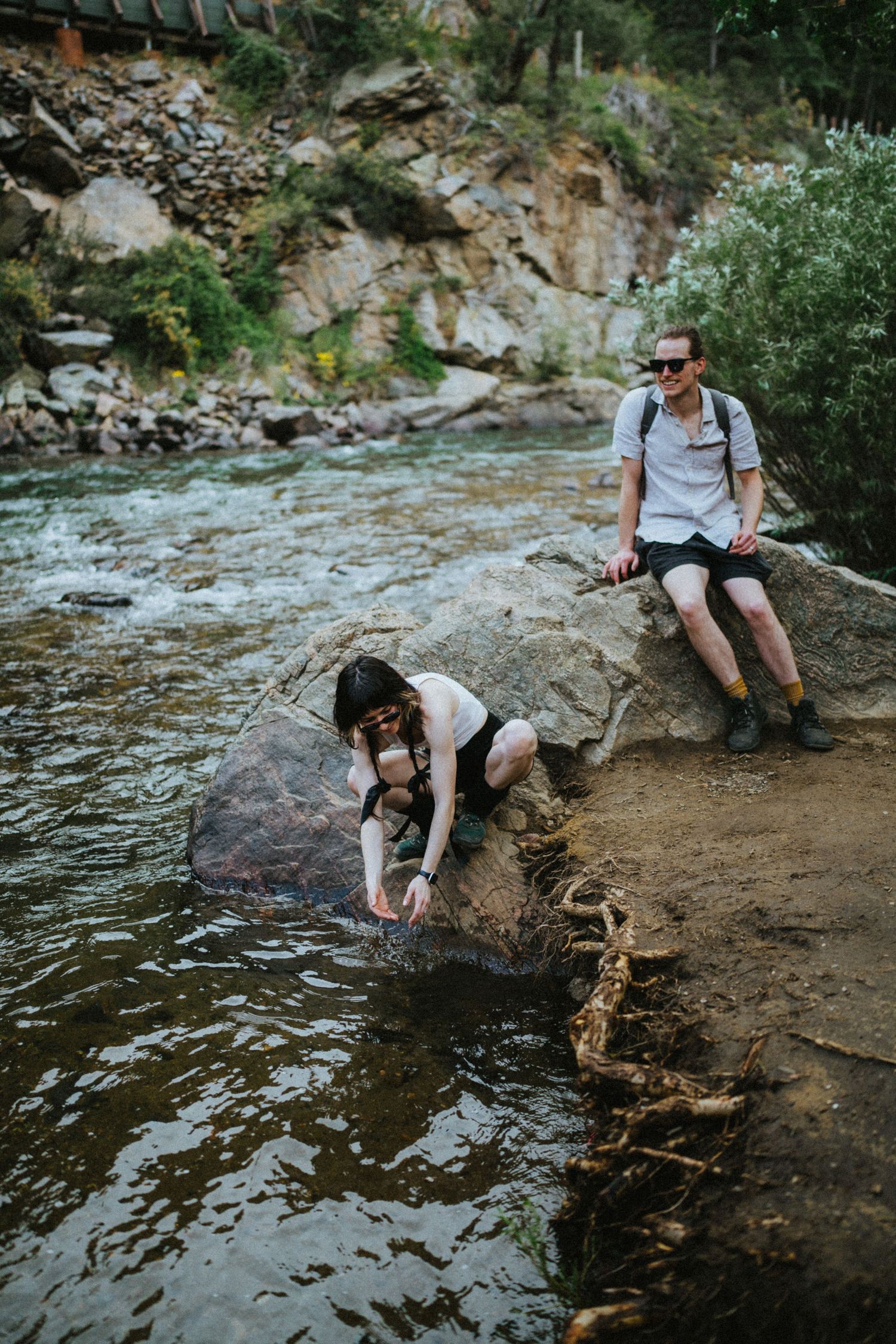 Woman touching water at rocky riverbank while man sits on boulder with backpack in mountain canyon.