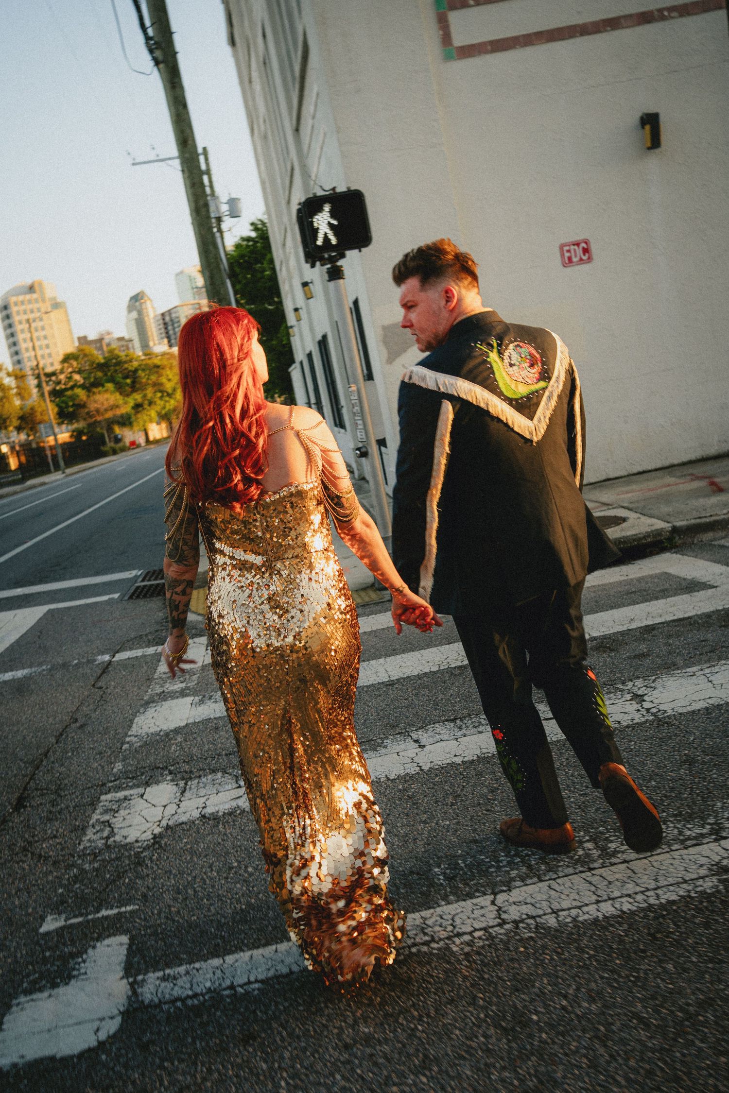 Couple crossing a street, woman in gold sequin gown with red hair, man in black embroidered jacket.