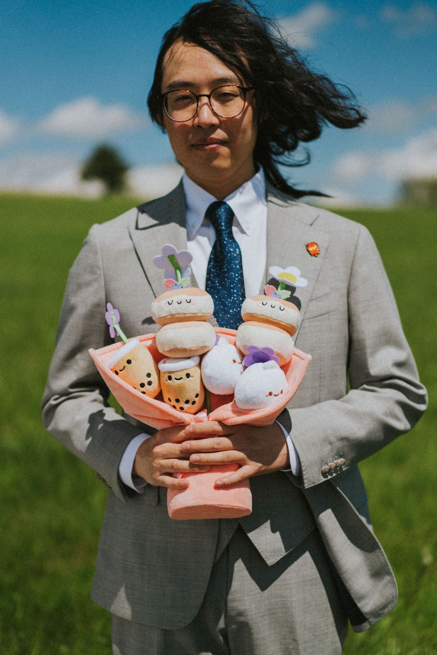 A person in a gray suit holds an assortment of plush toys while standing in a grassy field on a sunny day.