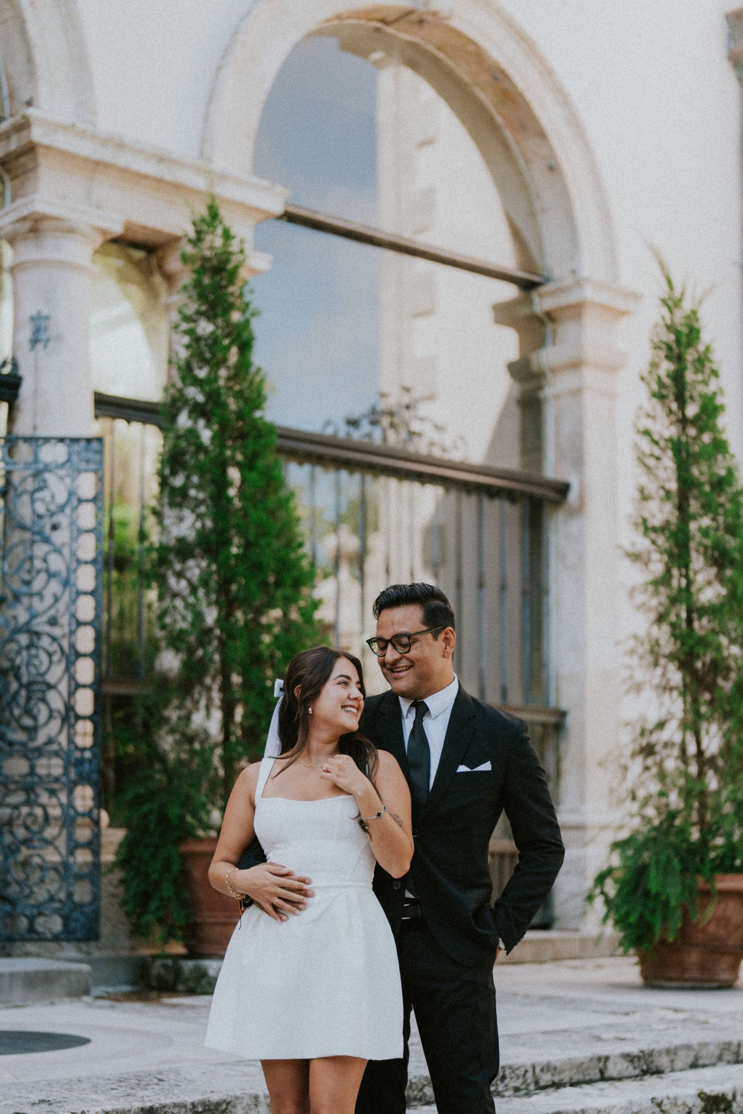 Couple in wedding attire smiling outdoors, bride in white mini dress with veil, groom in black suit, elegant stone building backdrop.