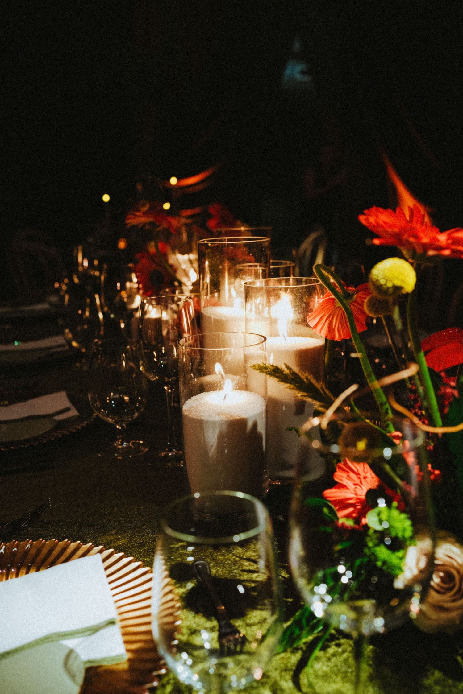 Elegant candlelit dinner table with glowing pillar candles, red gerbera daisies, and crystal glassware for a romantic event.
