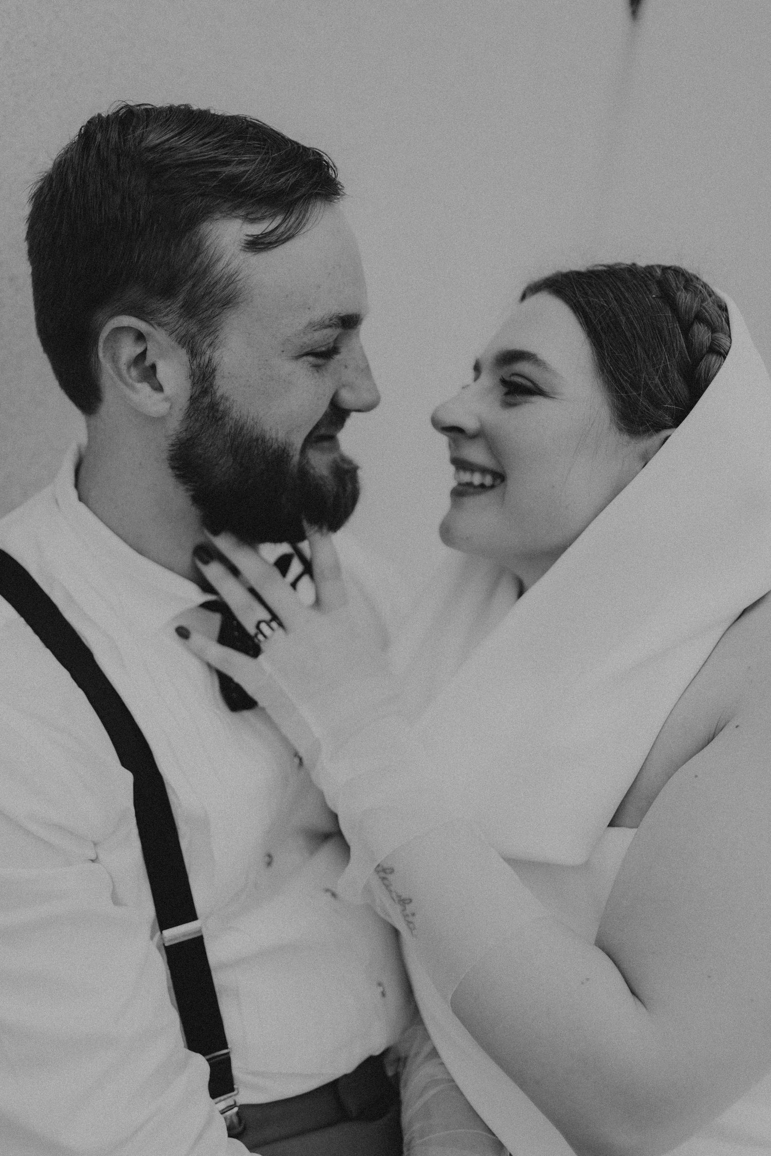 Black and white photo of smiling bride and bearded groom in suspenders gazing at each other lovingly.
