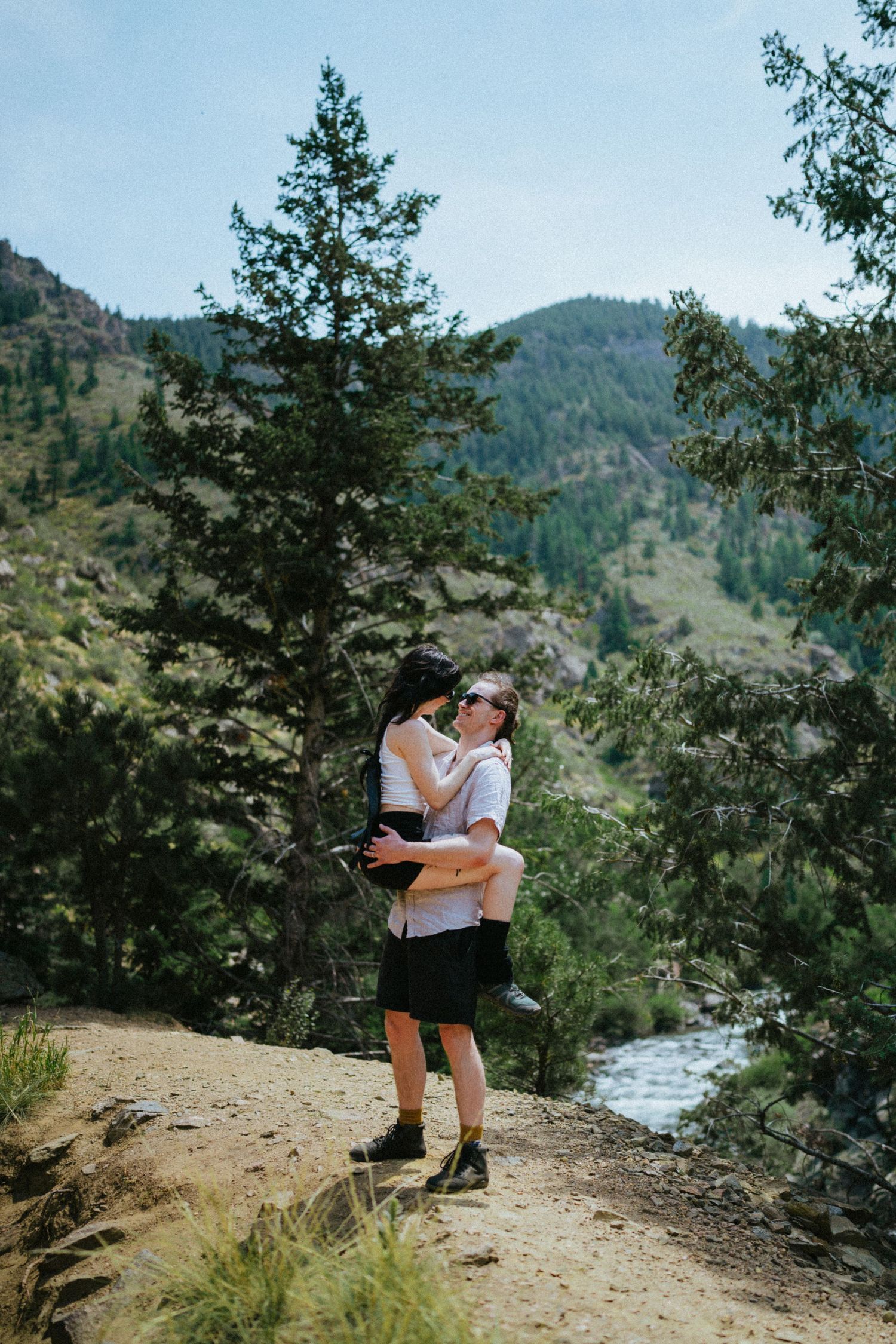 Couple embracing on a mountain hiking trail surrounded by pine trees and a scenic river valley.
