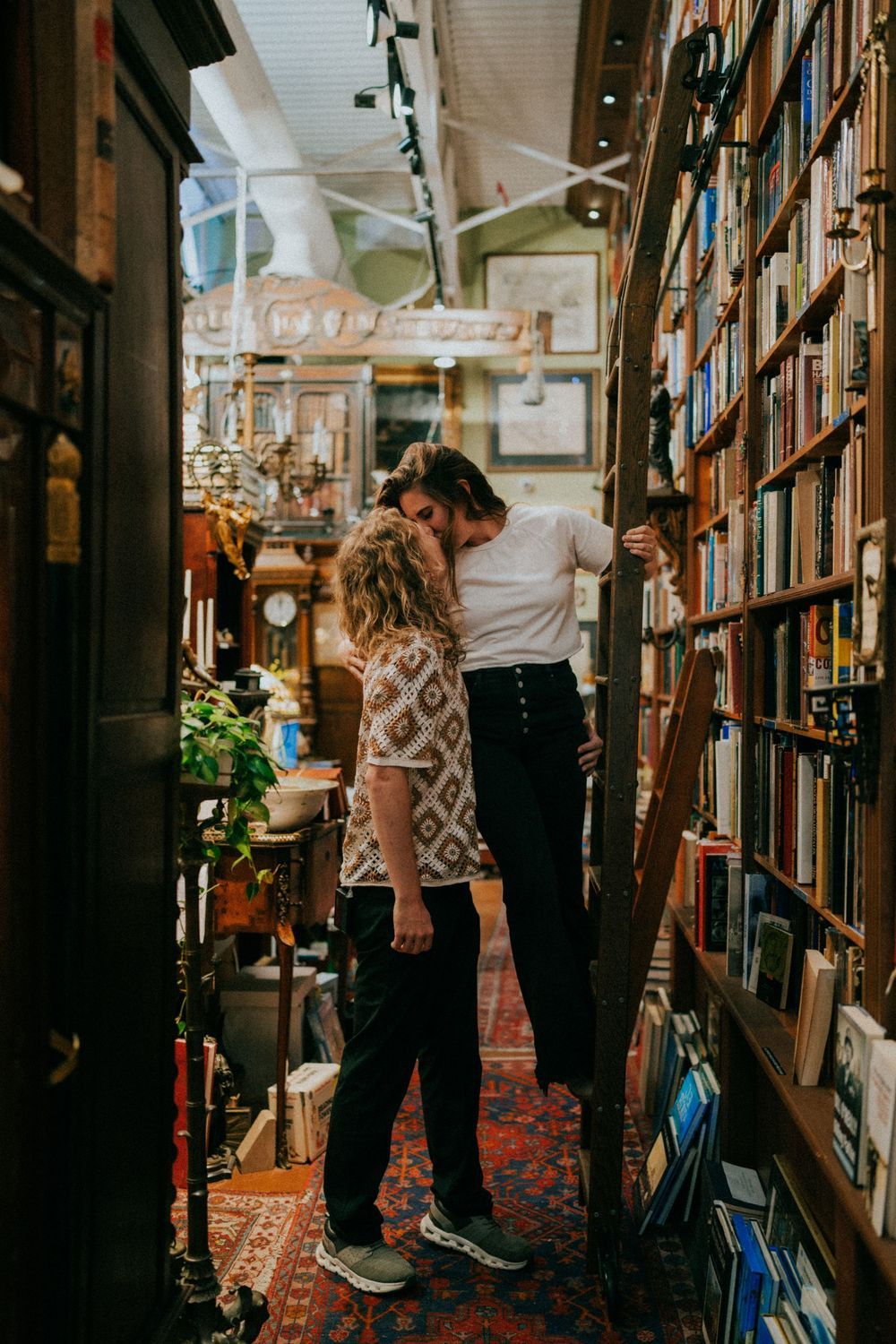A couple sharing romantic moments between bookshelves in a cozy library setting.