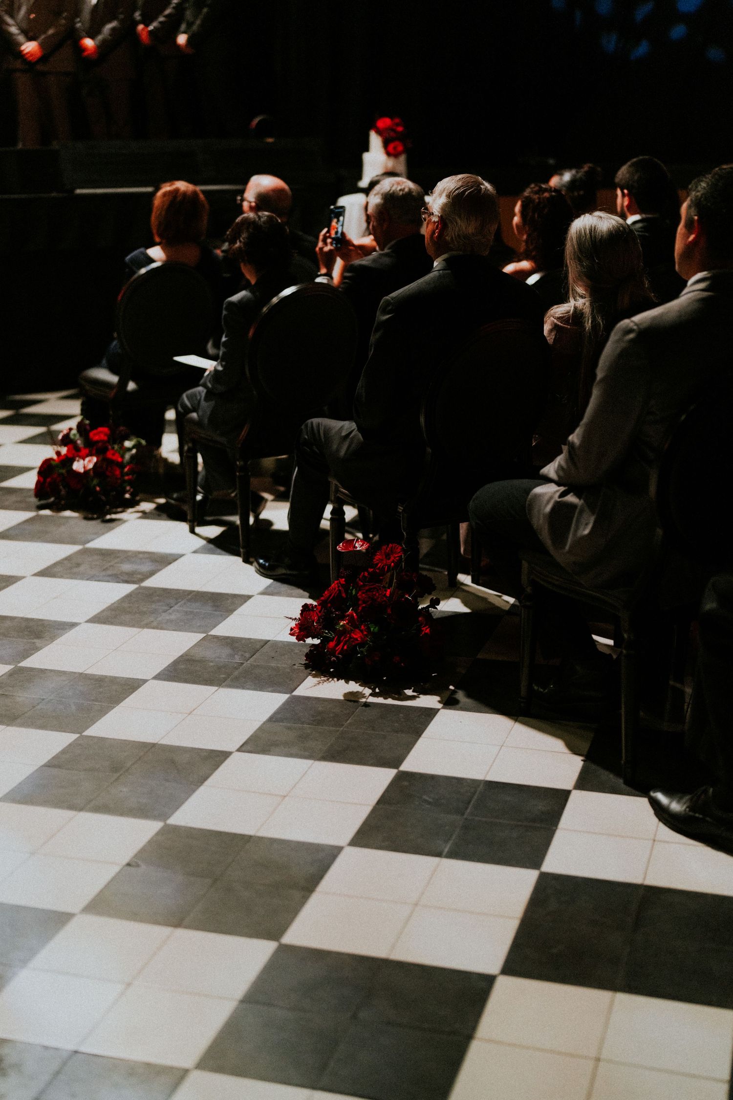 Wedding guests seated on chairs along a black and white checkered floor with red floral arrangements.