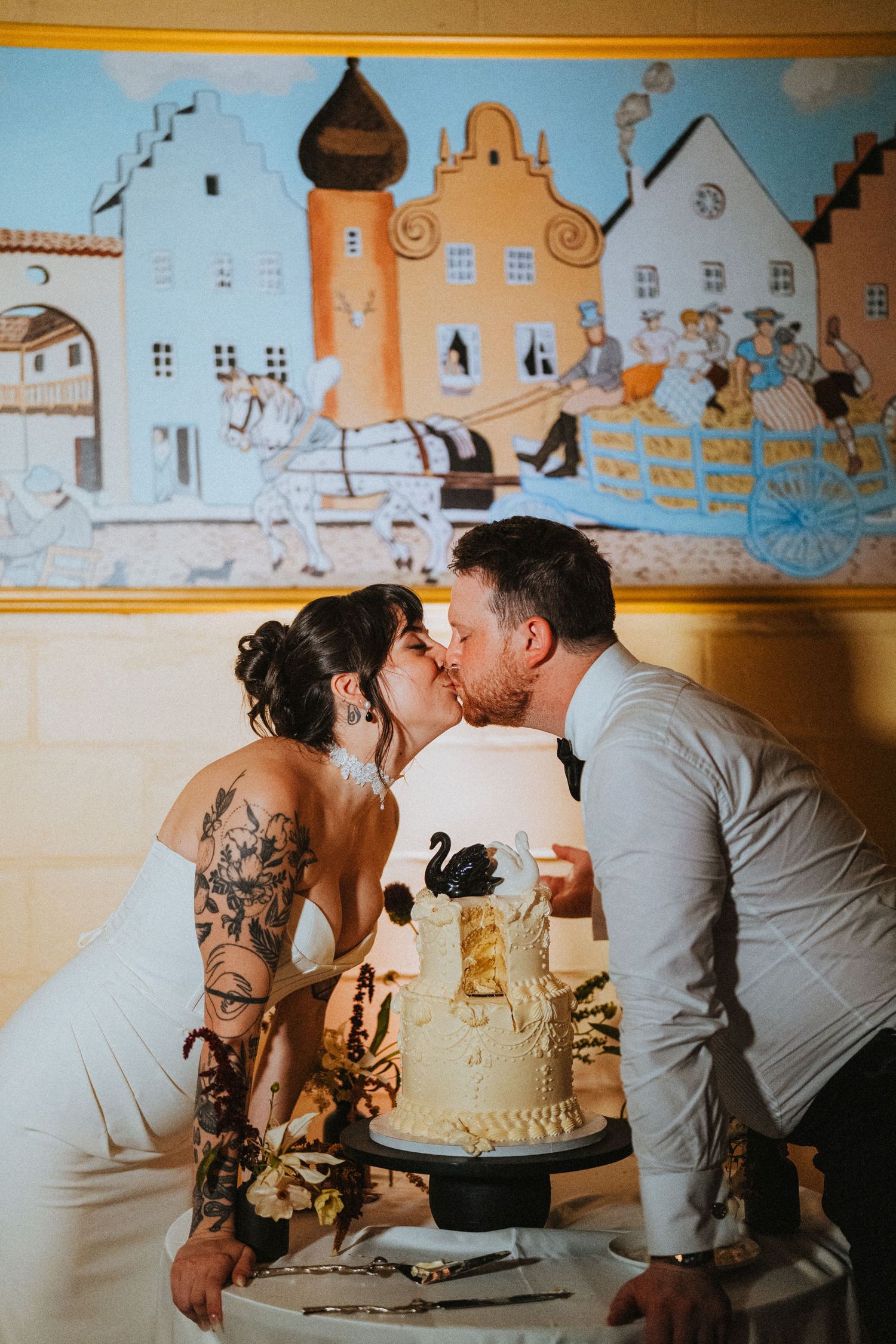 Tattooed bride and groom share a kiss behind a elegant black and gold wedding cake at their reception.