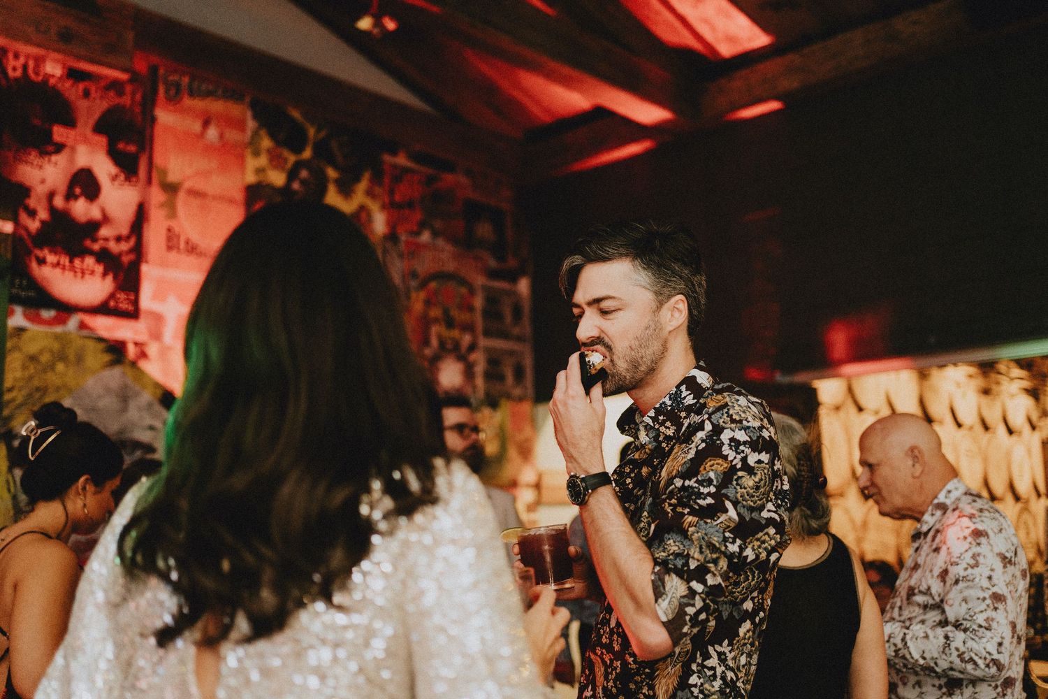 Man in floral shirt at a lively party venue with colorful decorations and posters in the background.