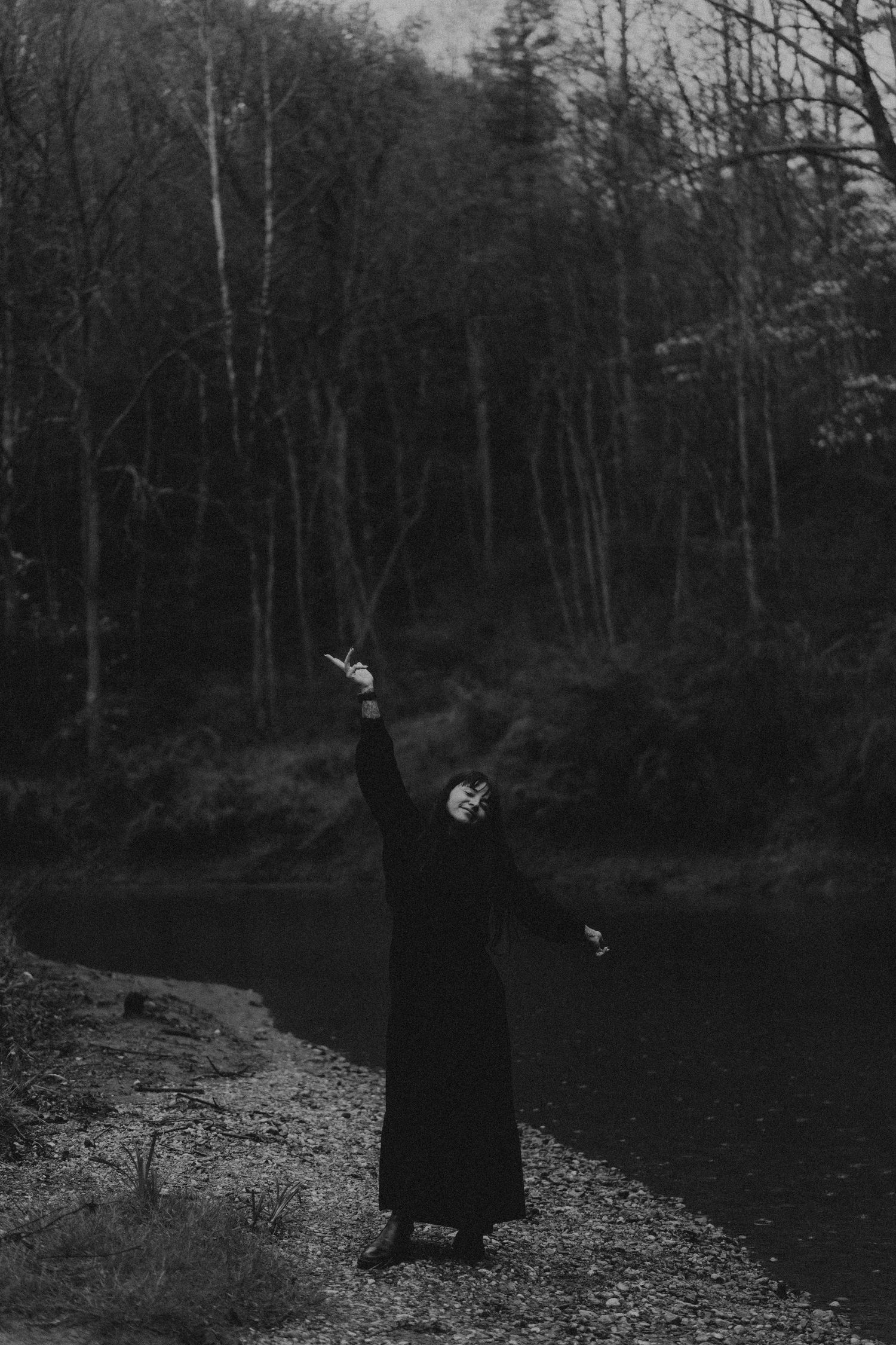 A figure in dark clothing stands on a rocky riverbank in a moody black and white forest setting.