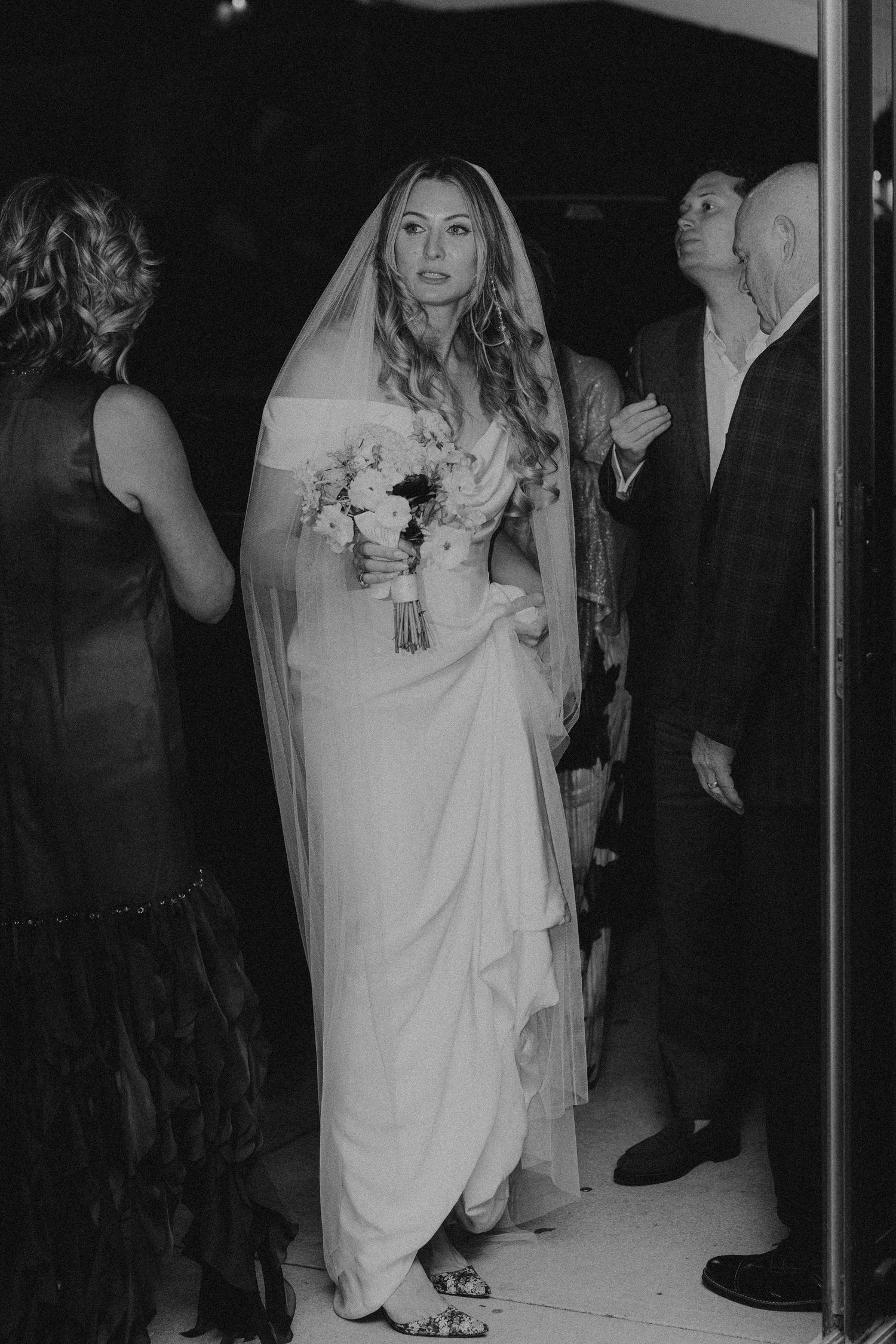 Black and white photo of a bride in a flowing white gown and veil holding a bouquet at her wedding ceremony.