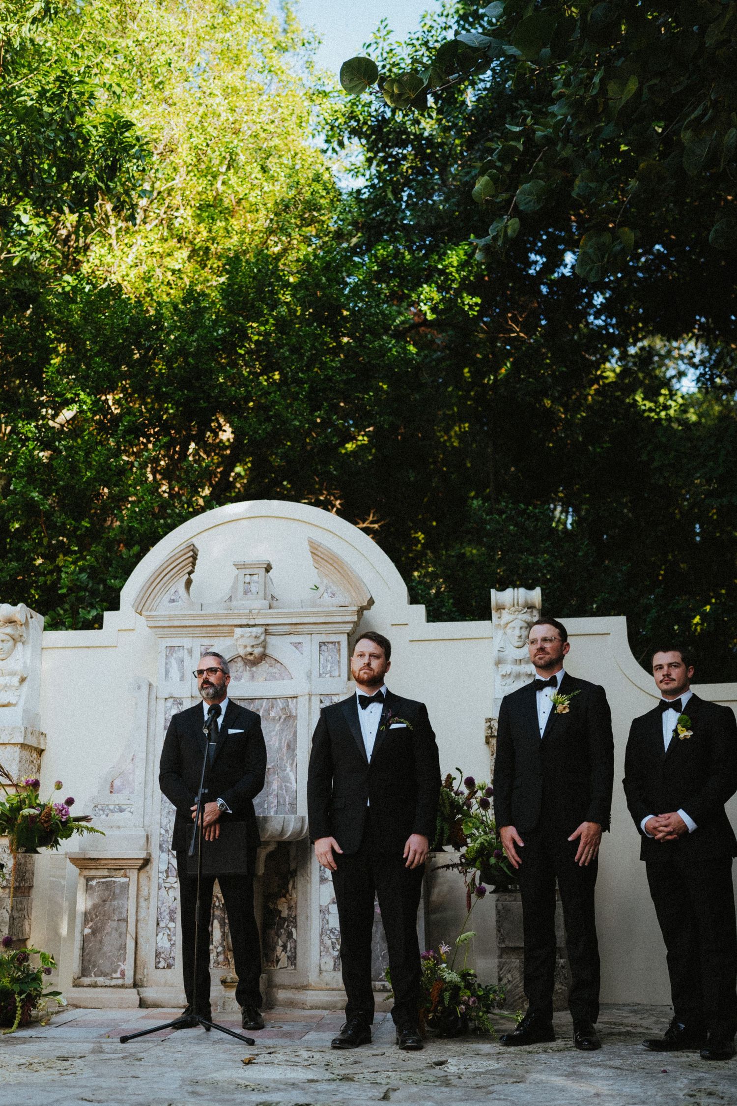 Four groomsmen in black tuxedos standing before ornate white stone archway surrounded by lush green trees.
