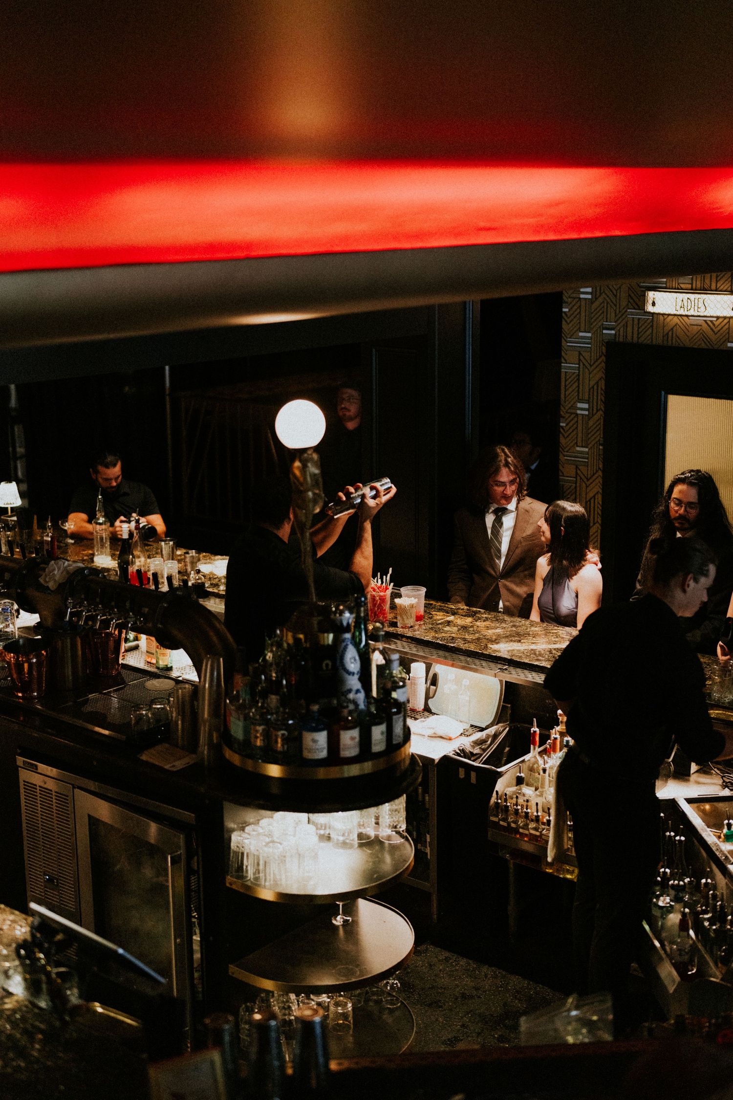 Dimly lit upscale bar with bartenders serving guests under red ambient lighting and a glowing pendant light.