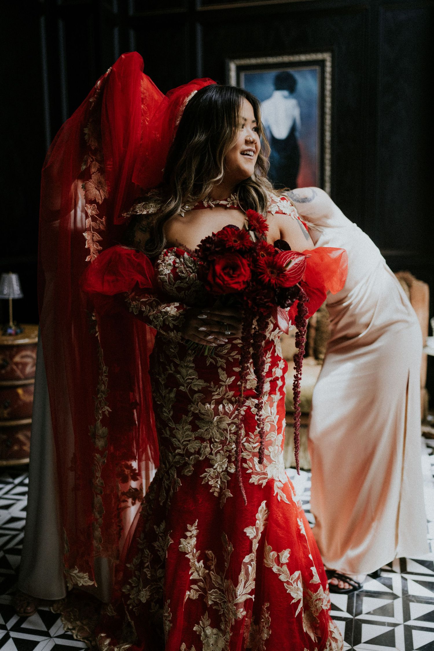 Bride in red floral off-shoulder gown with red veil holds bouquet of red roses in elegant dark interior.