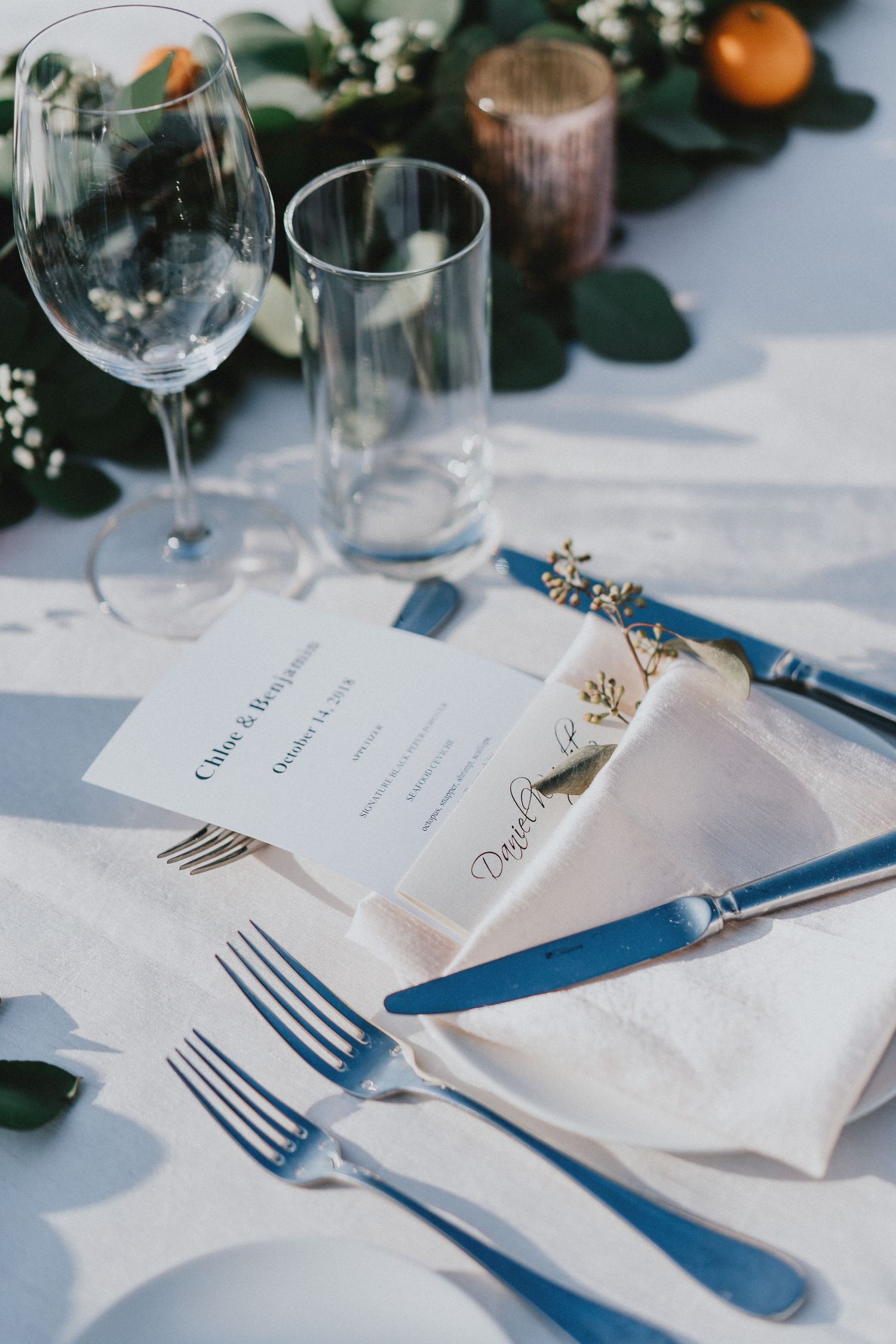 Elegant wedding table setting with blue cutlery, white napkins, wine glasses, and floral greenery centerpiece.
