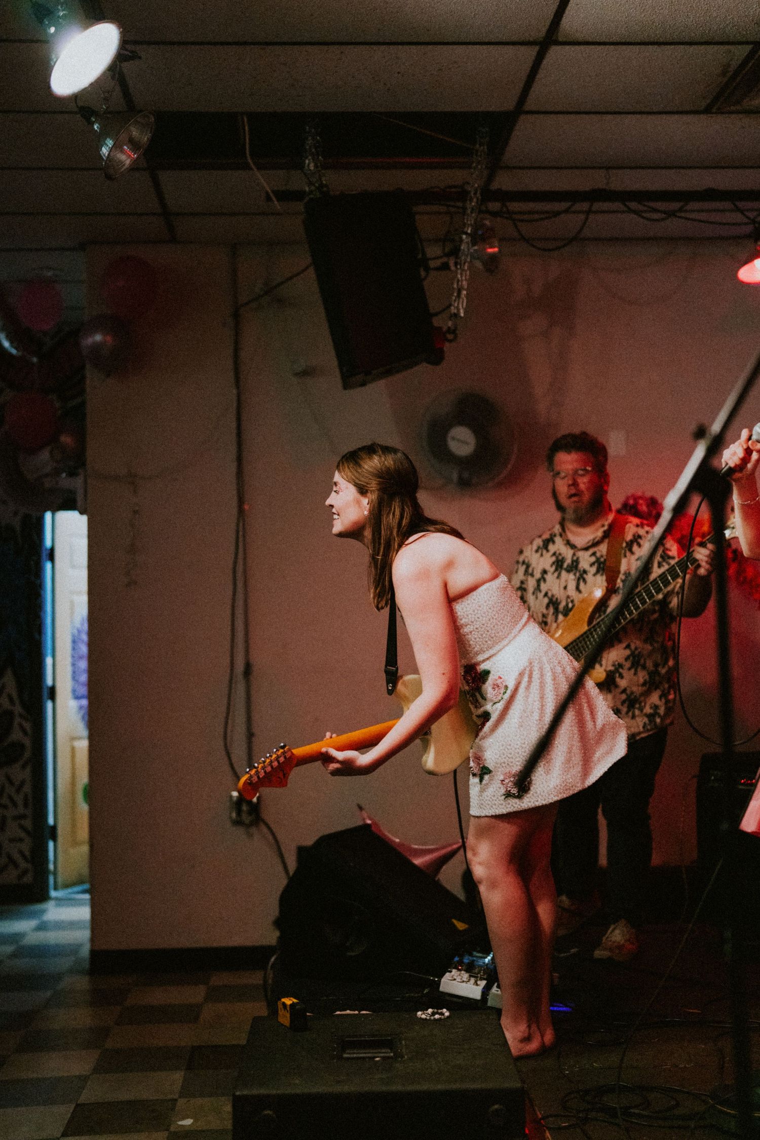 Two musicians performing on stage, woman in white dress playing guitar, man in floral shirt with bass guitar.