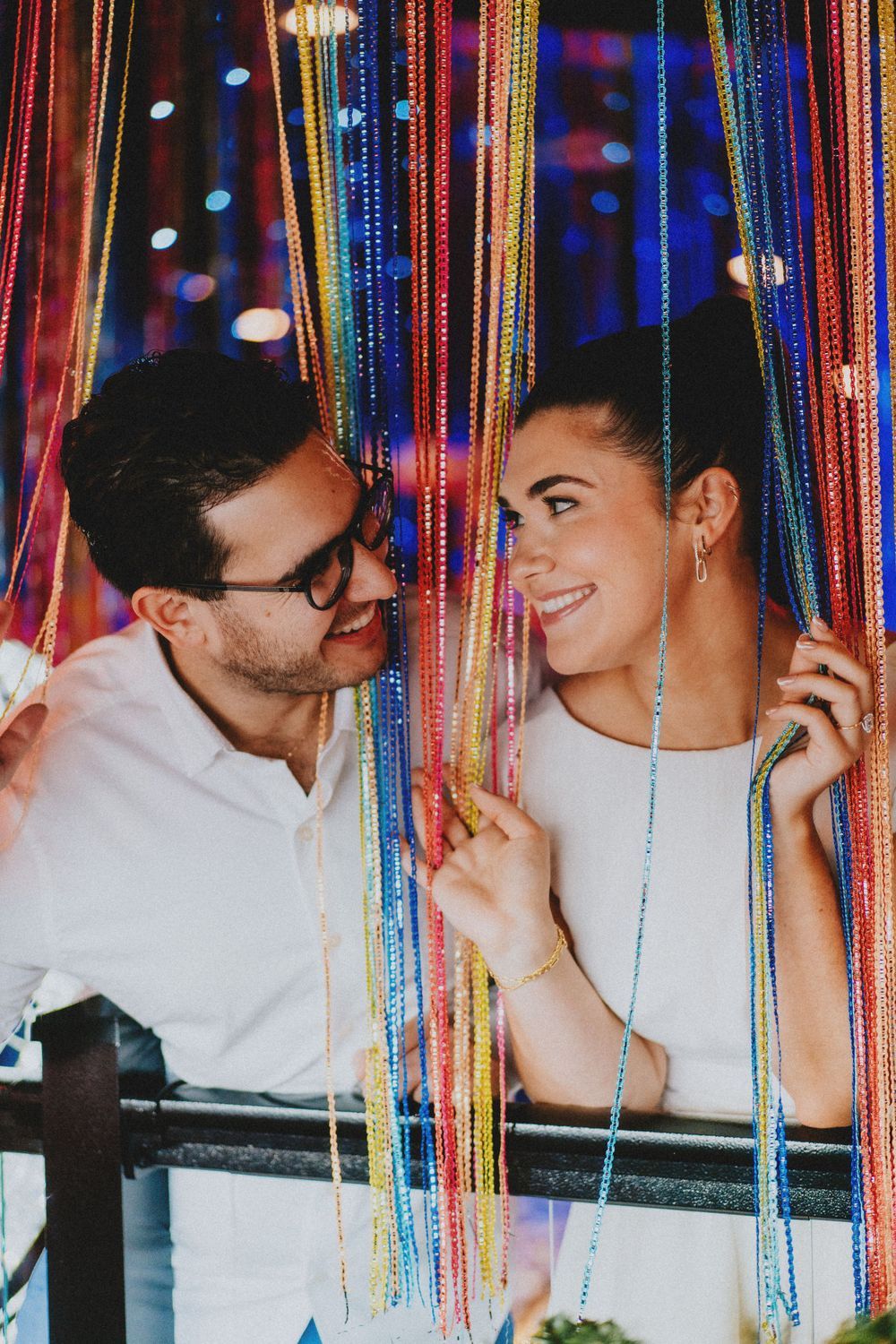 A couple sharing tender moments behind colorful hanging beads with bokeh lighting effects.