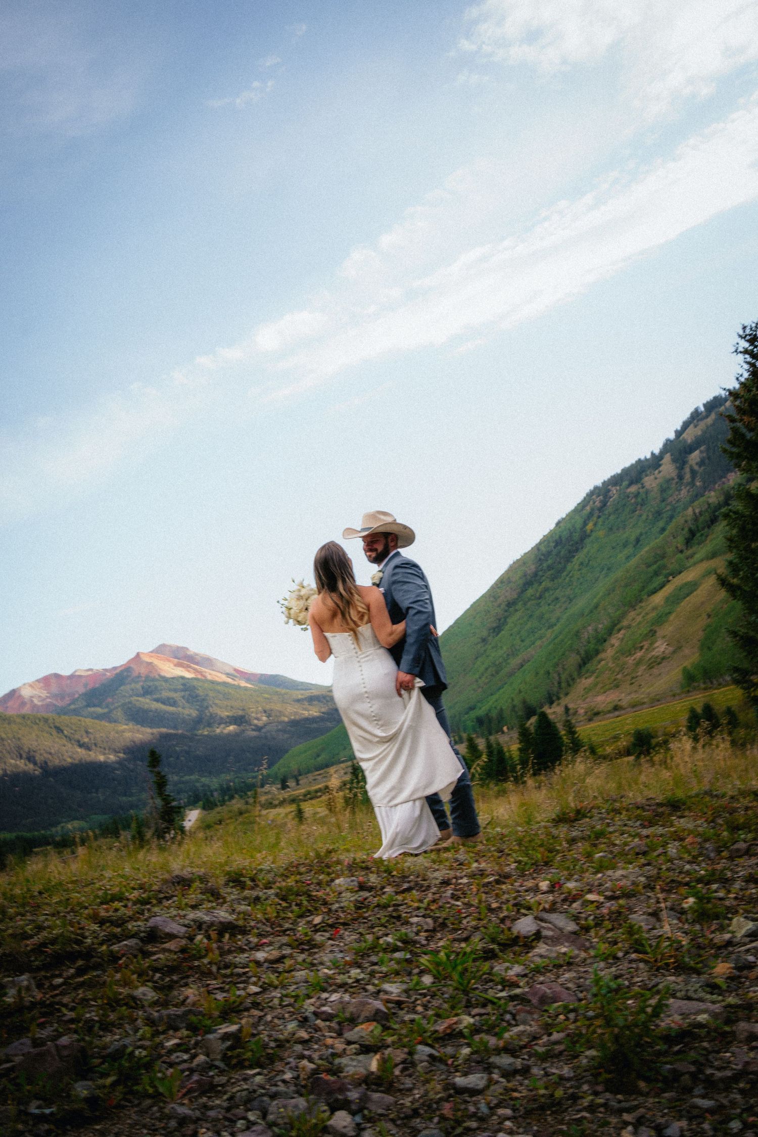A bride and groom embrace on a rocky mountain trail surrounded by lush green hills and a clear blue sky.