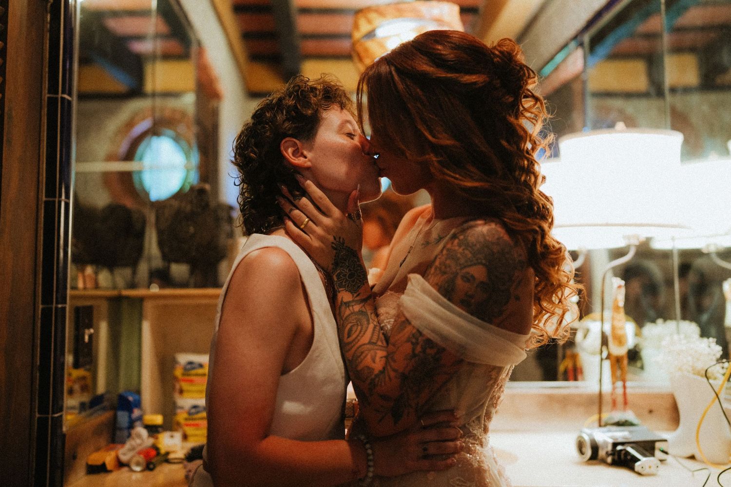 Two people sharing a romantic kiss in a warmly lit room, one wearing a white dress with tattoos.