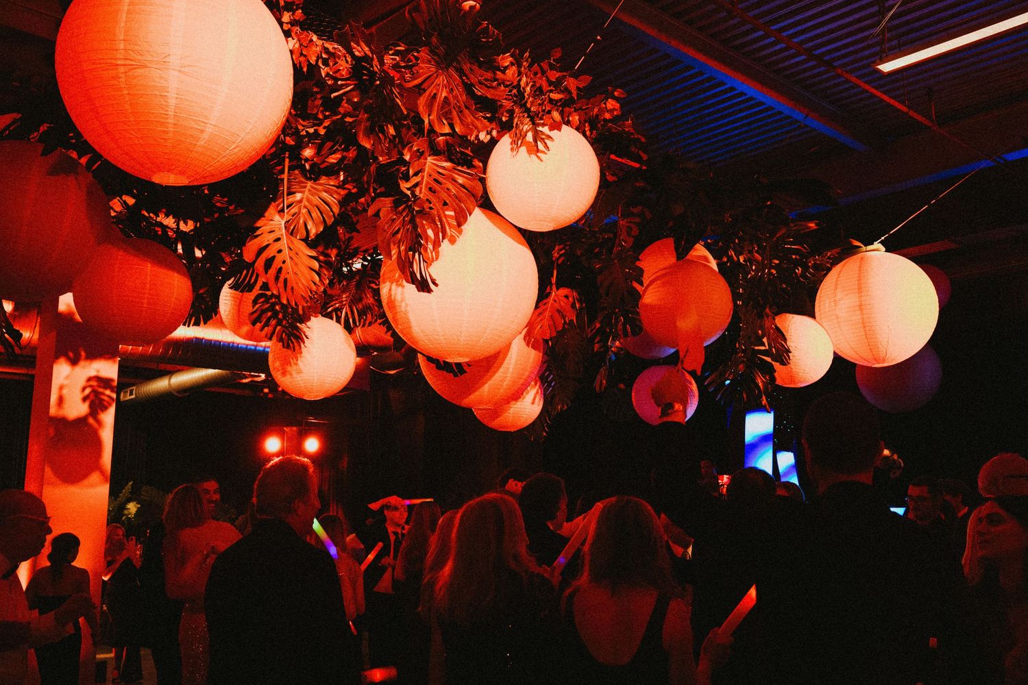 Glowing paper lanterns hanging above a crowd at an indoor evening event with warm red and orange lighting.