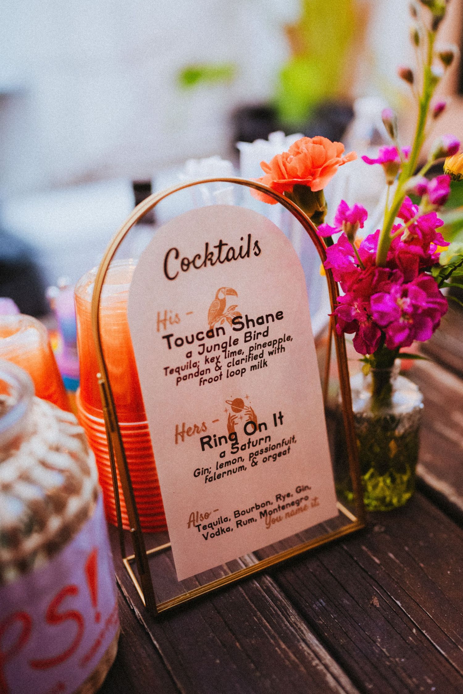 Gold-framed cocktail menu sign with his-and-hers drinks, flanked by pink flowers and orange straws at a wedding reception.