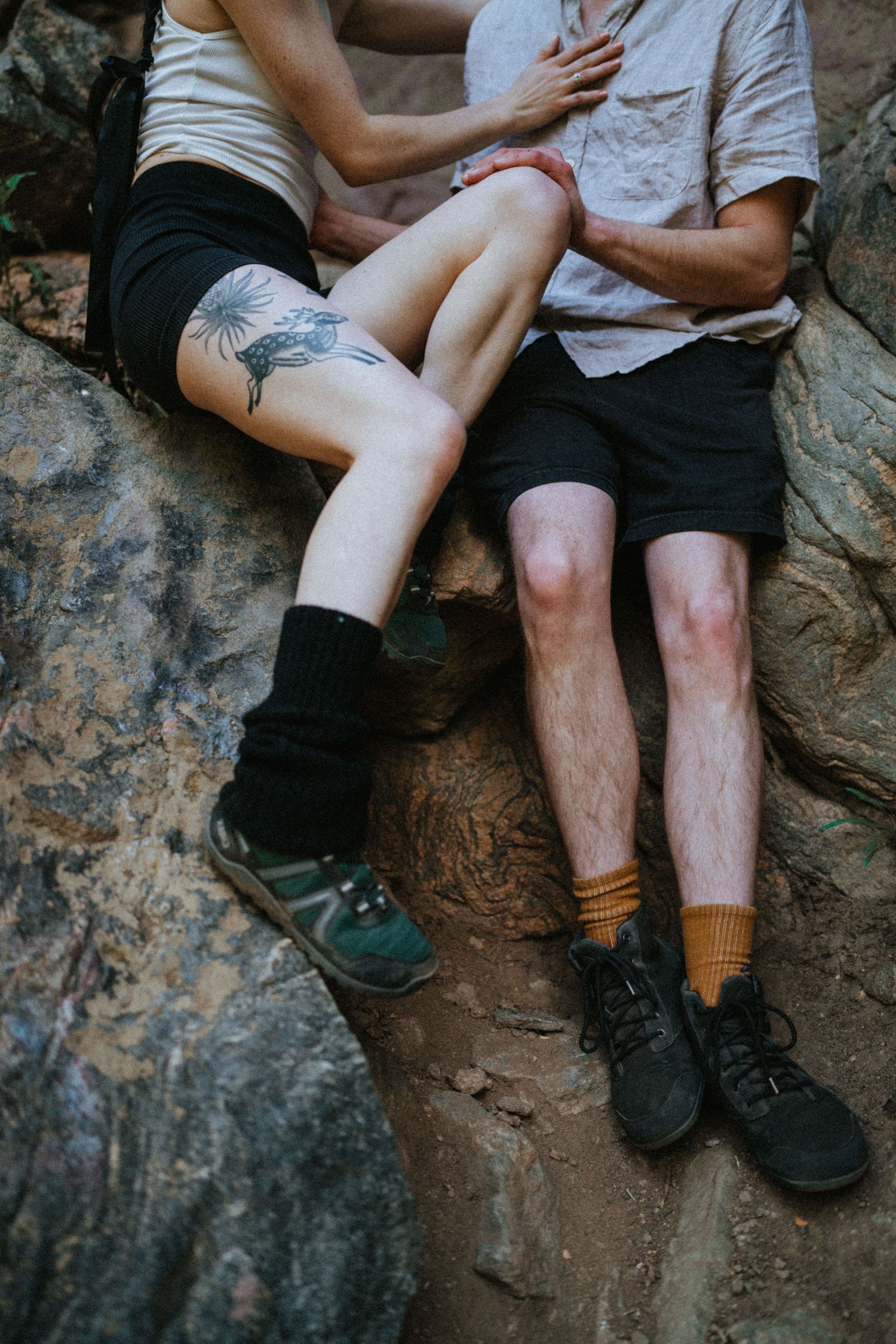 Couple sitting together on rocky terrain, one with floral leg tattoo, wearing hiking shorts and colorful socks.
