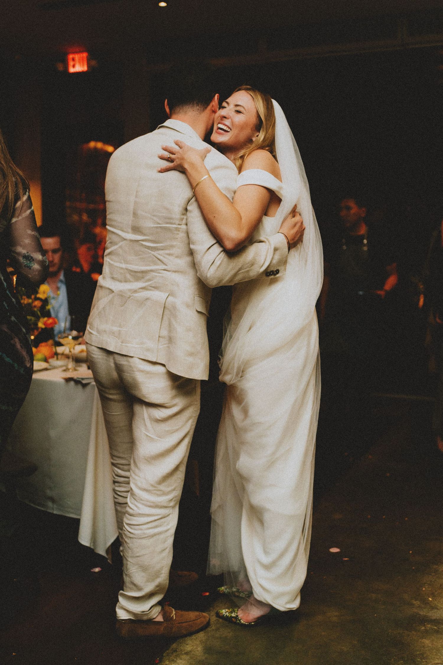 Bride in white off-shoulder gown sharing a joyful embrace with a guest at wedding reception.