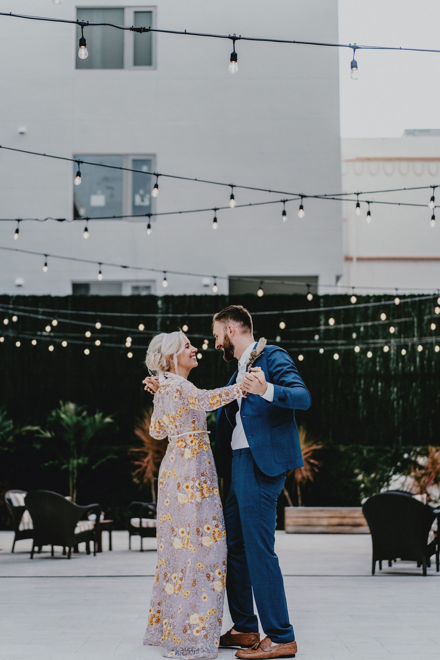 Couple sharing first dance on outdoor patio with string lights at wedding reception, woman in floral gown, man in blue suit.