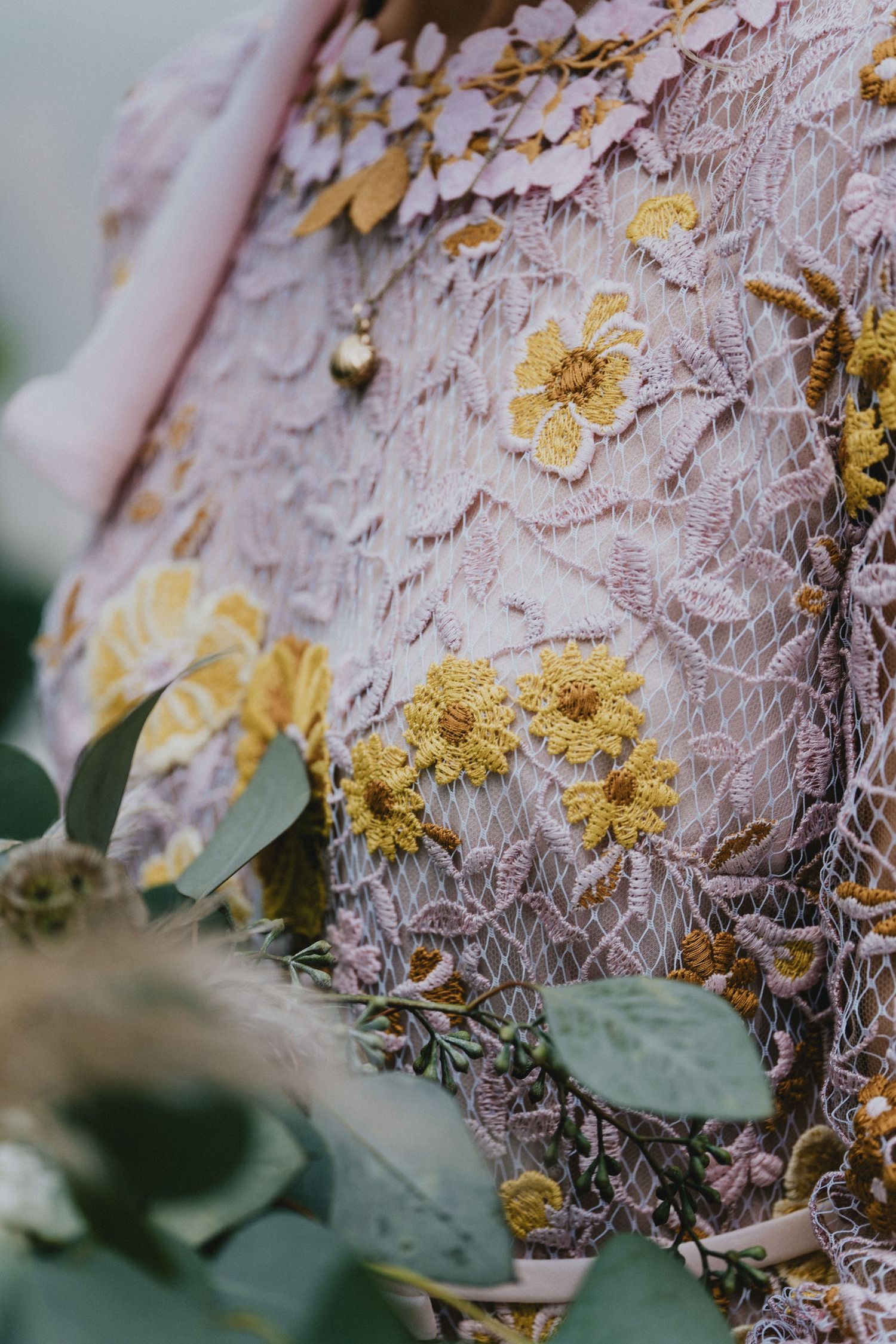 Close-up of elegant pink lace fabric with intricate yellow floral embroidery, surrounded by soft green eucalyptus leaves.