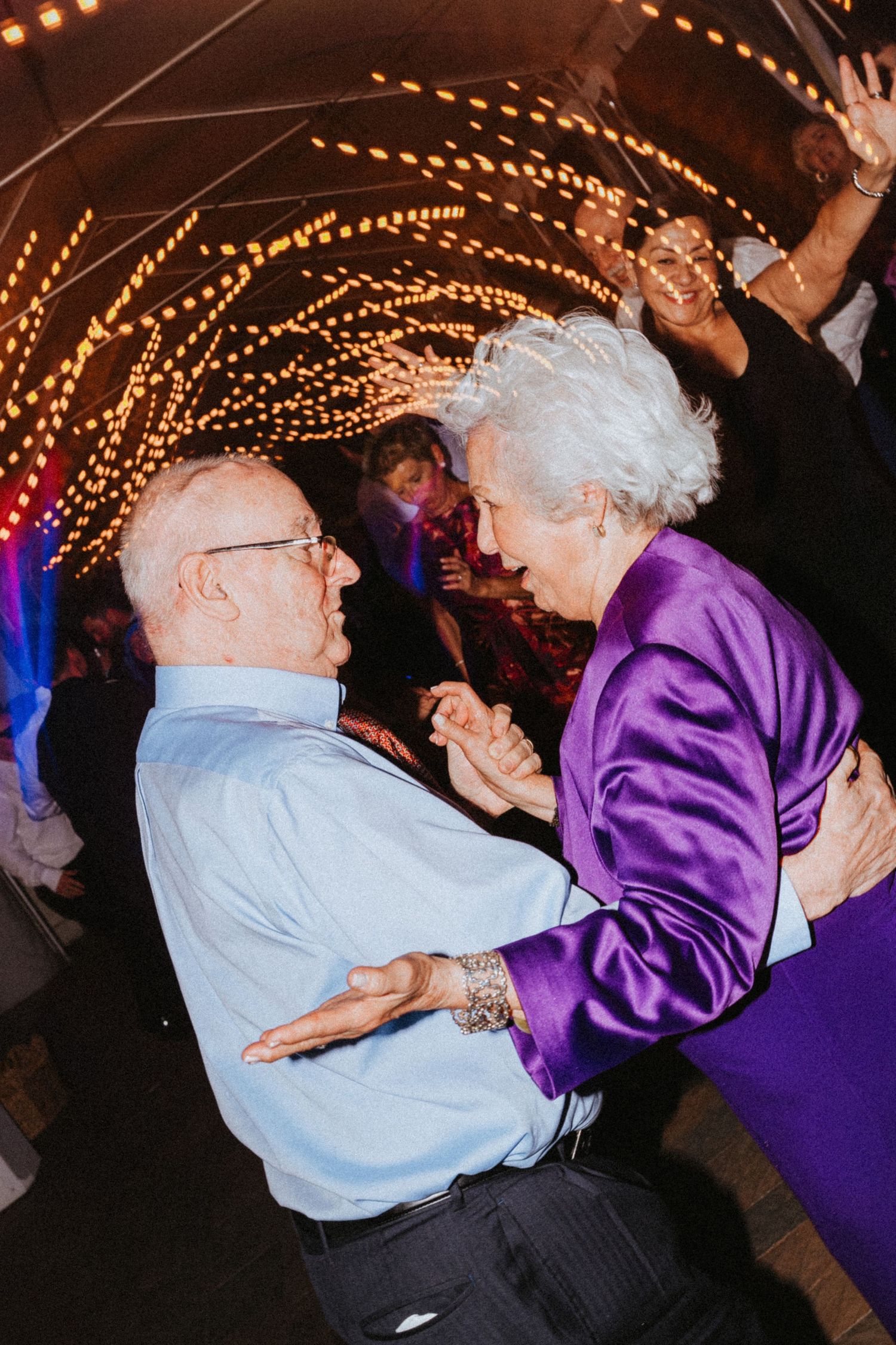 Elderly couple dancing joyfully together under glowing lights at an elegant wedding reception celebration.
