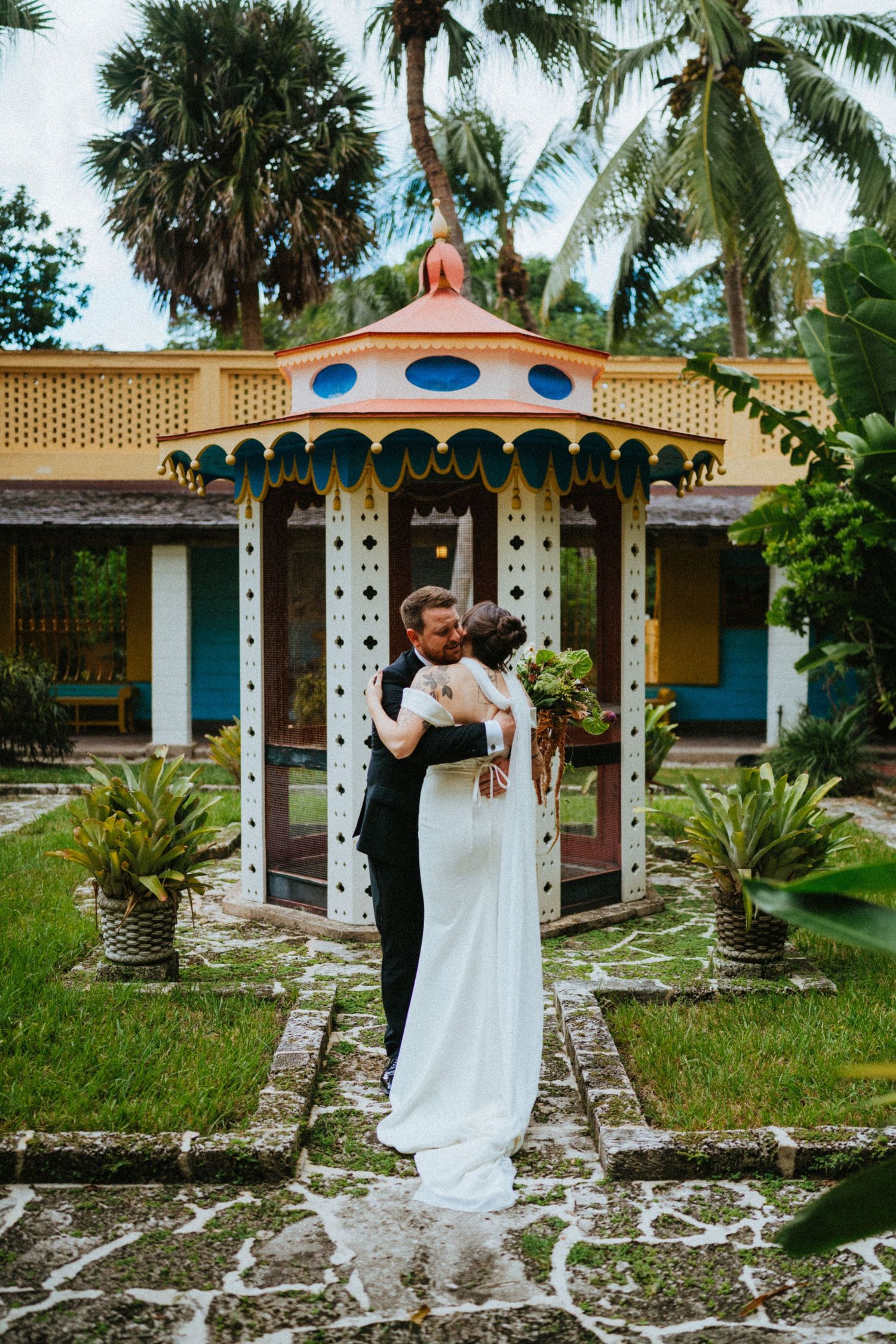 Bride and groom share first kiss in front of colorful tropical gazebo surrounded by lush greenery.