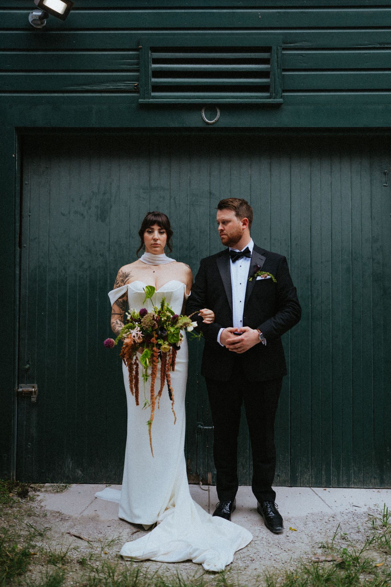 Bride in white gown holding wild bouquet stands with groom in black tuxedo before green barn doors.