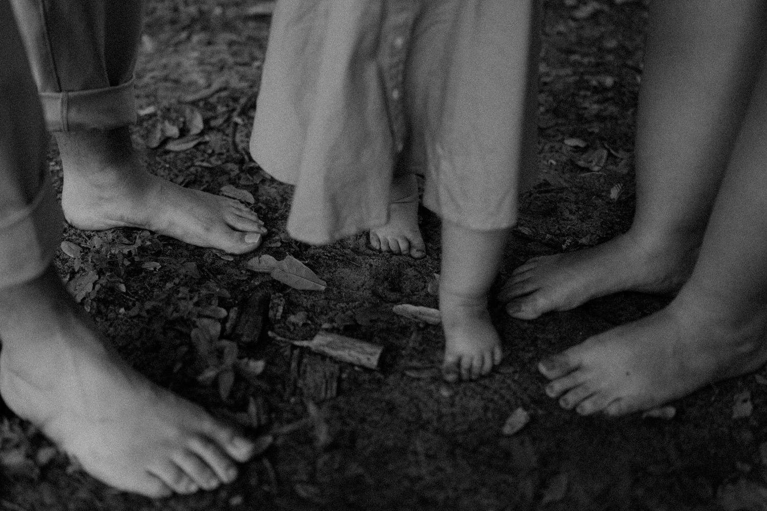 Black and white photo of barefoot family standing together on dirt ground, showing feet of adults and small child.