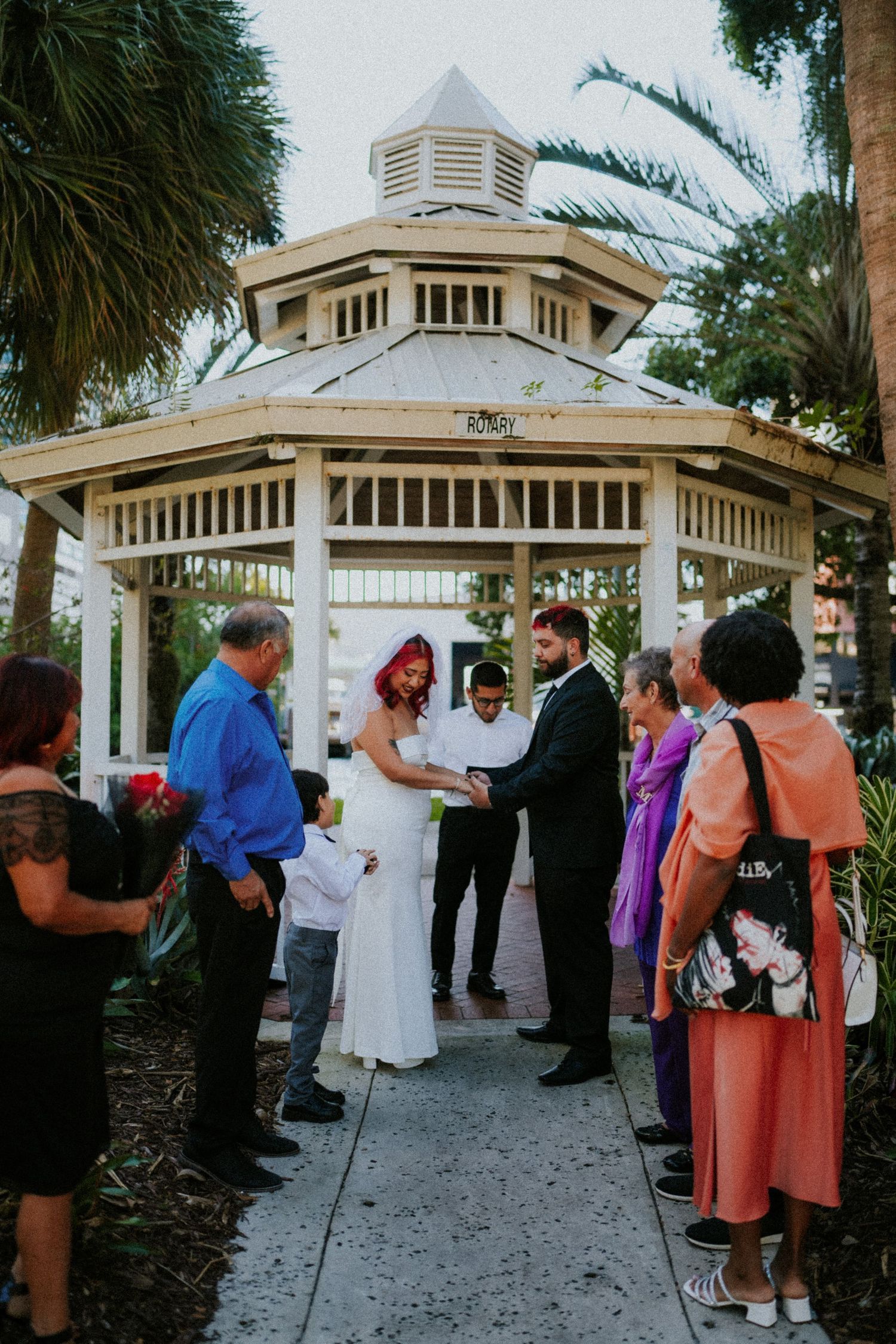 Bride and groom exchange vows under a white gazebo surrounded by guests during an outdoor wedding ceremony.
