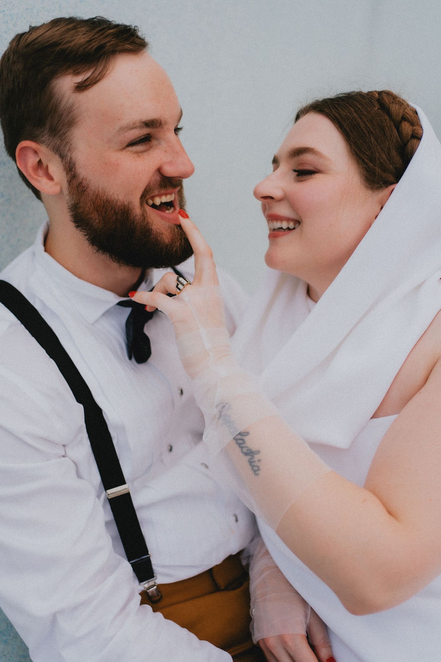 Laughing bride in white veil playfully touches bearded groom's face during wedding portraits.