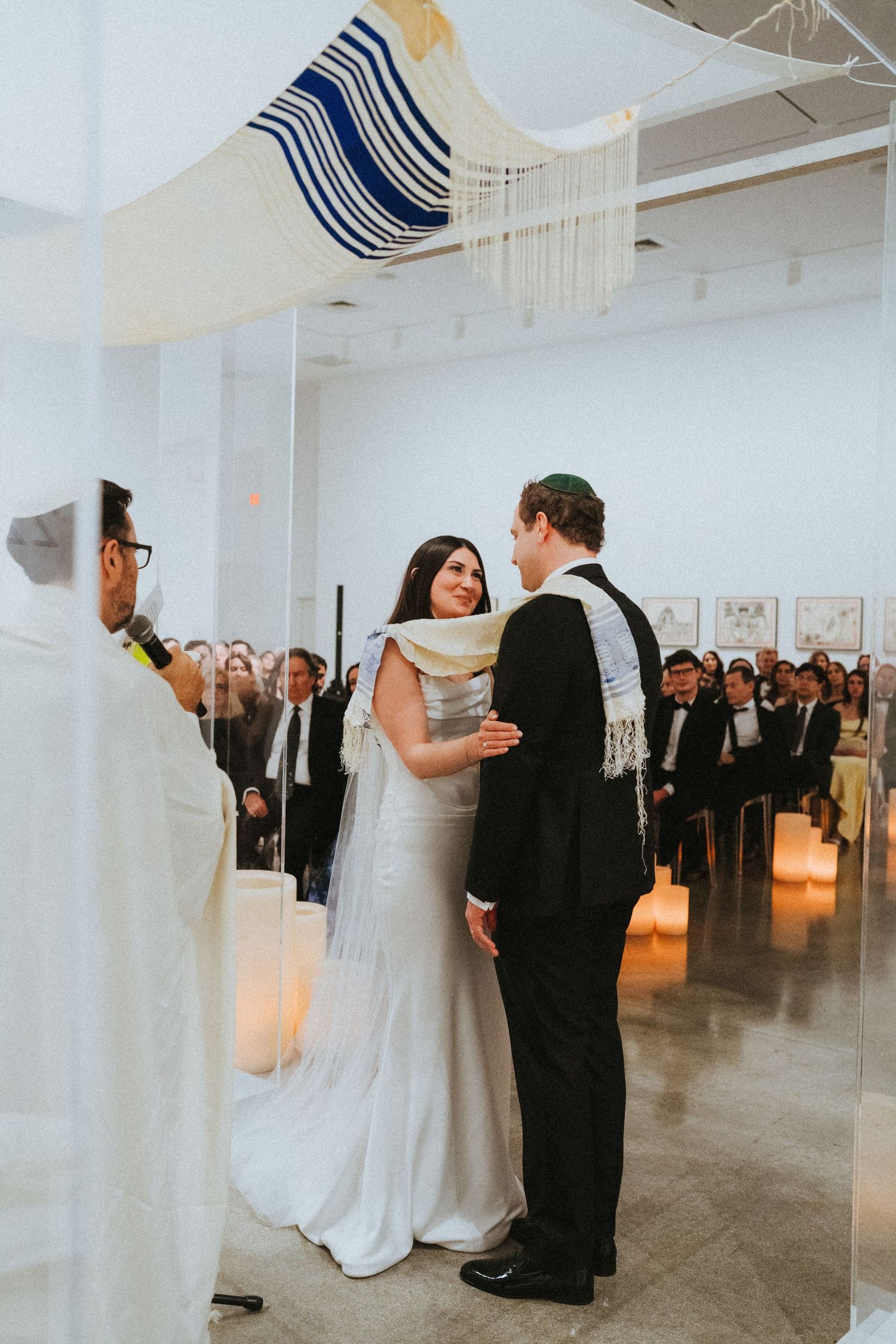 Bride and groom exchange vows under a tallit chuppah during Jewish wedding ceremony in modern art gallery.