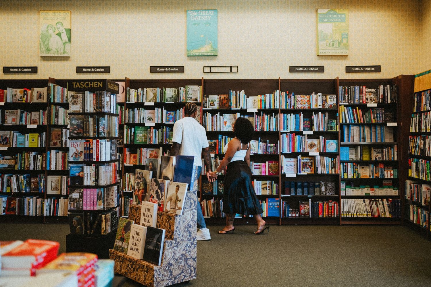 Two people browsing books in a well-stocked bookstore with floor-to-ceiling shelves filled with colorful titles.