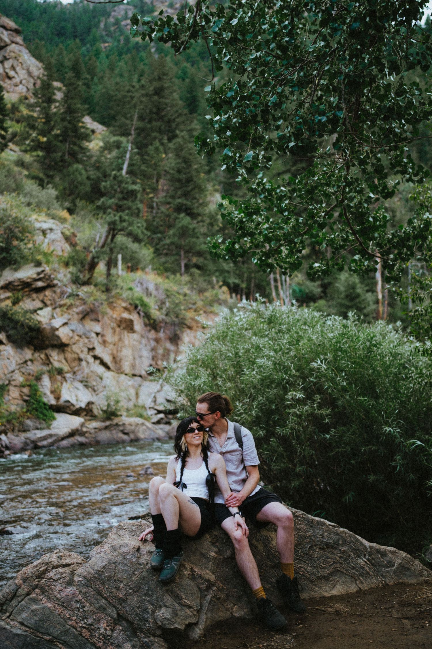 Couple sitting together on a boulder beside a mountain river surrounded by green trees and rocky cliffs.