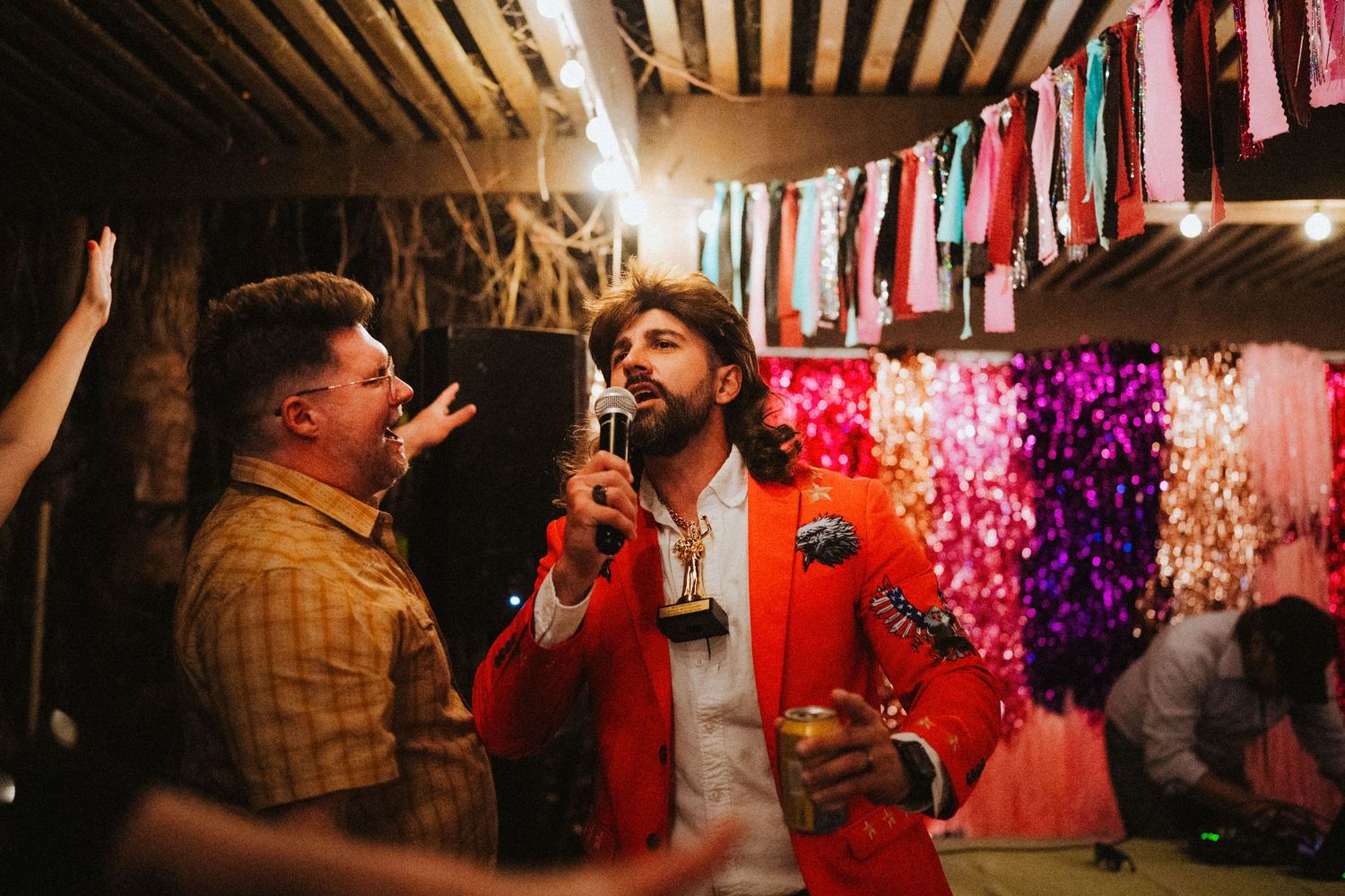 Two men celebrating at a colorful party venue with string lights and festive decorations, one wearing a red blazer.