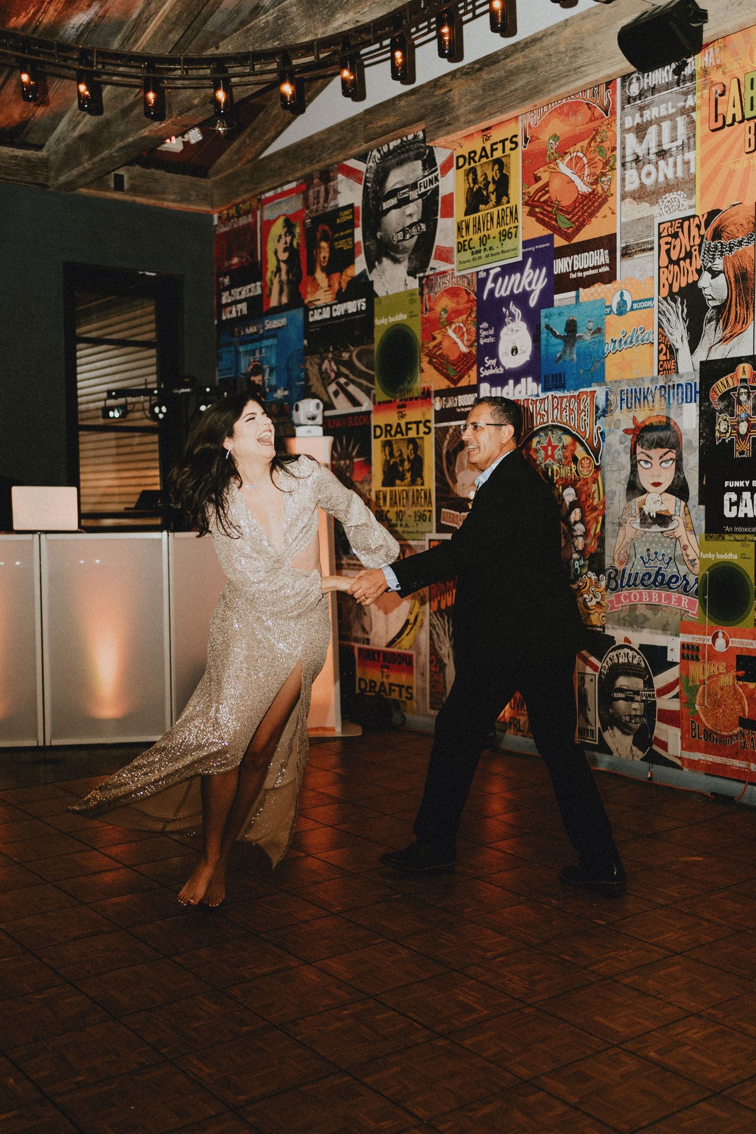 Bride in sequin dress dancing energetically with a guest in front of colorful music posters.