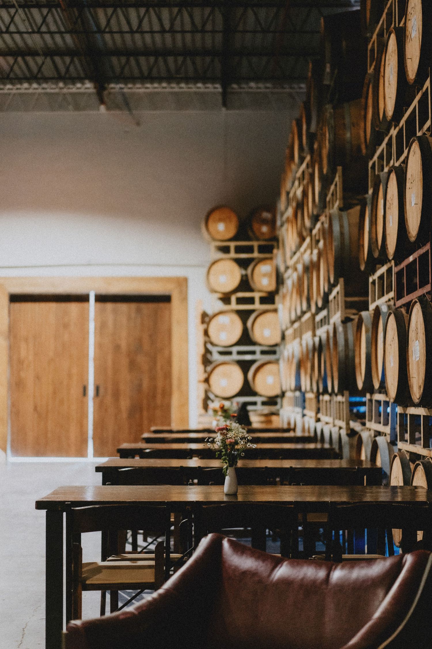 Rustic winery tasting room with leather chairs, wooden tables, and stacked oak barrels lining the wall.