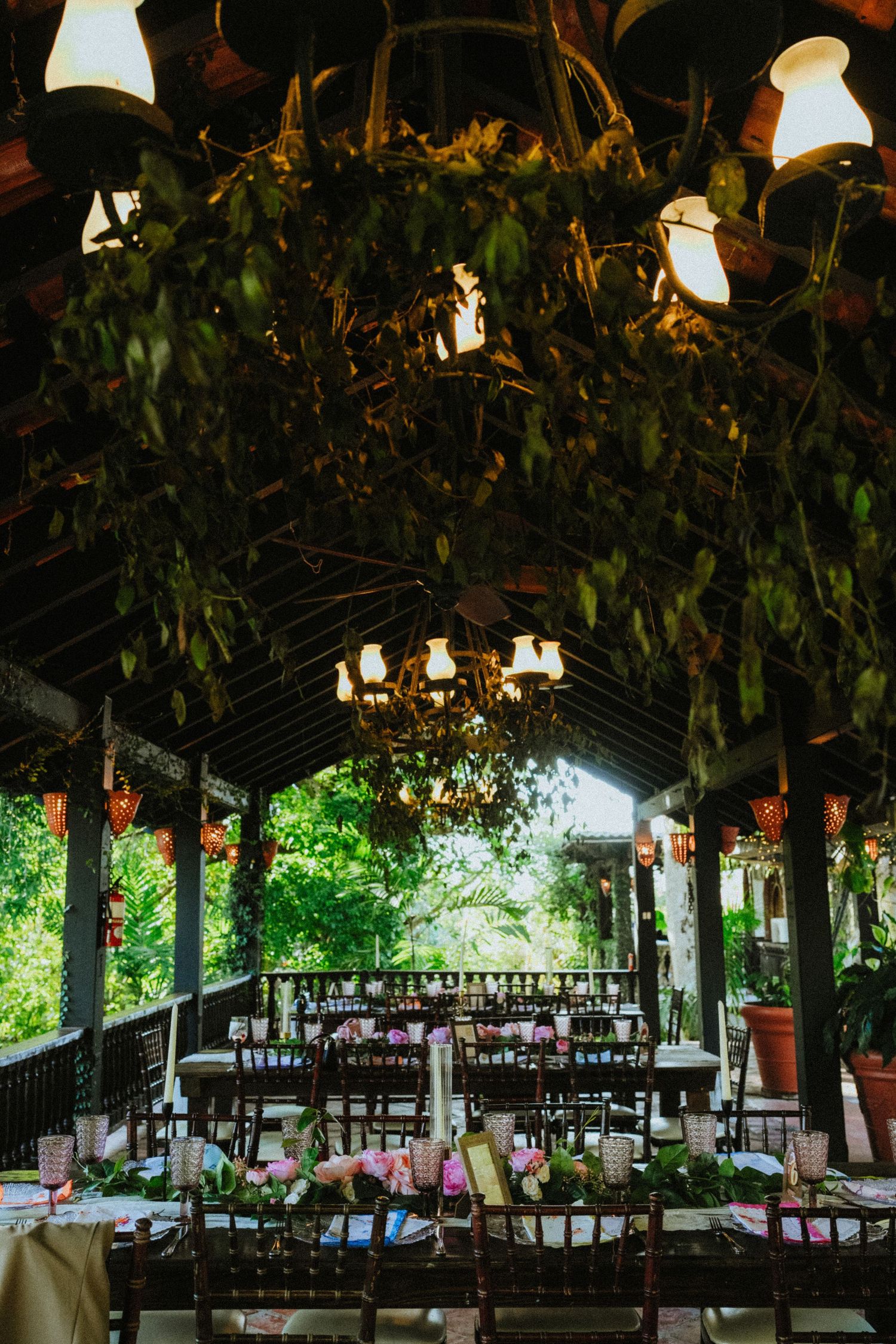 Rustic outdoor wedding reception venue with lush greenery ceiling, chandeliers, and elegantly set dining tables surrounded by nature.