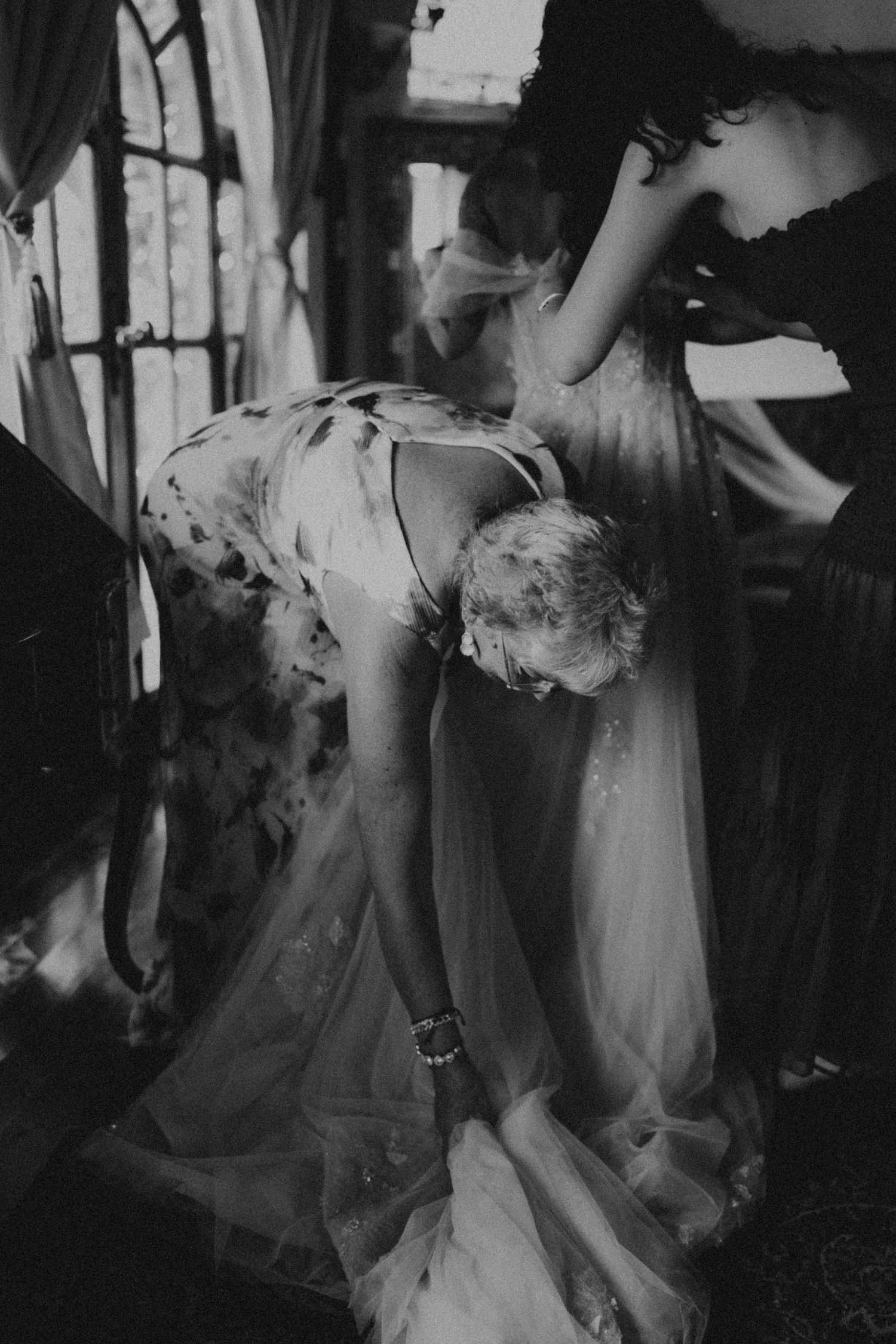 Black and white photo of a bride bending down adjusting her wedding dress train, assisted by a bridesmaid.