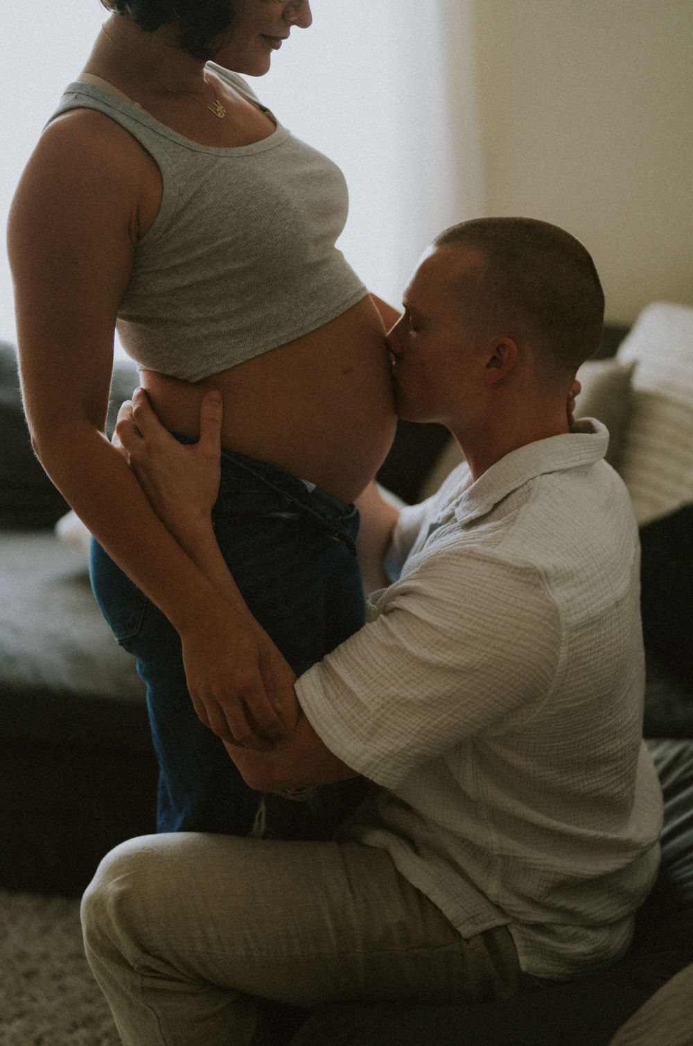 An intimate maternity photo shows a tender moment between expecting parents in a dimly lit room.
