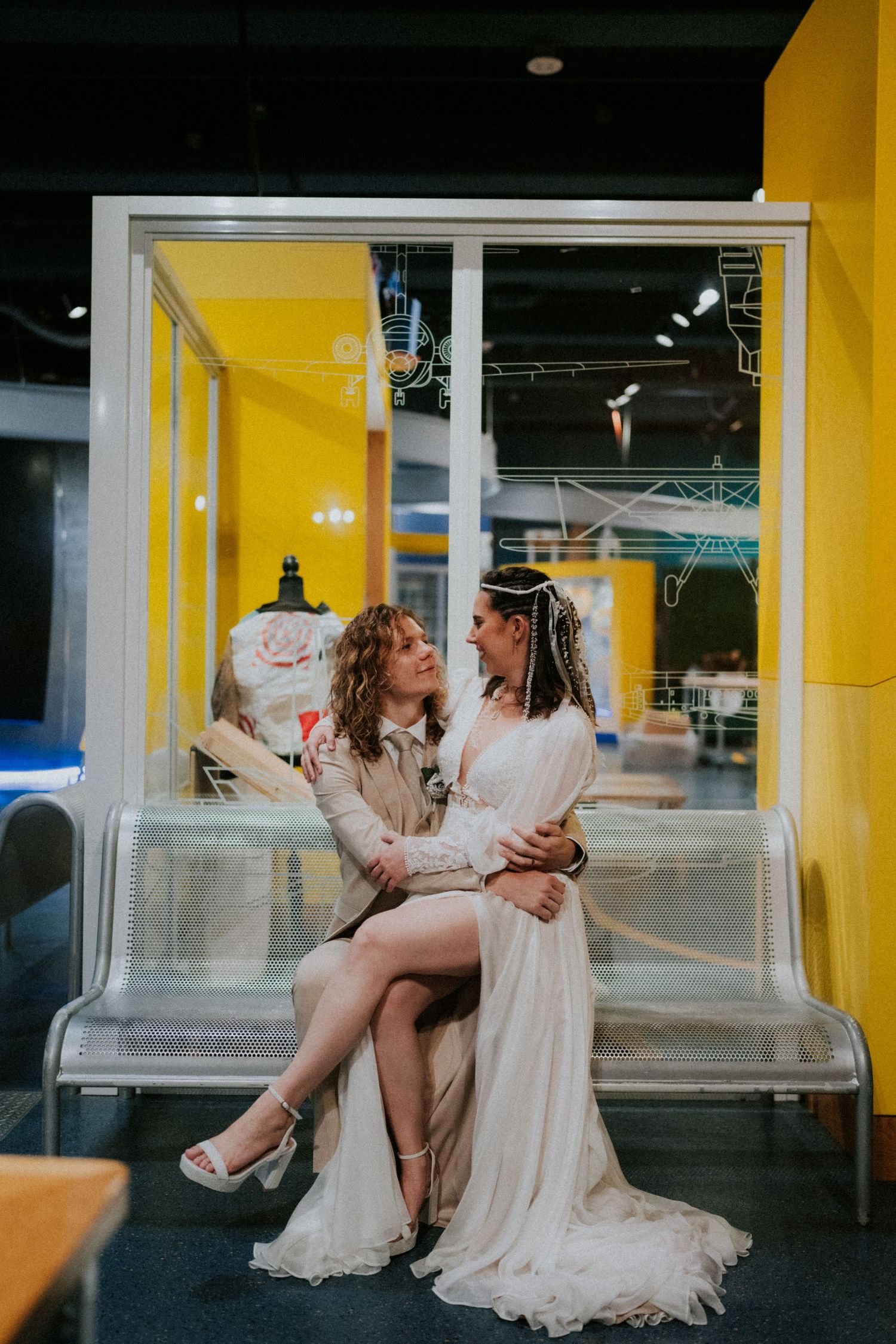 Two people in white dresses sit together on a modern bench with yellow walls in the background.