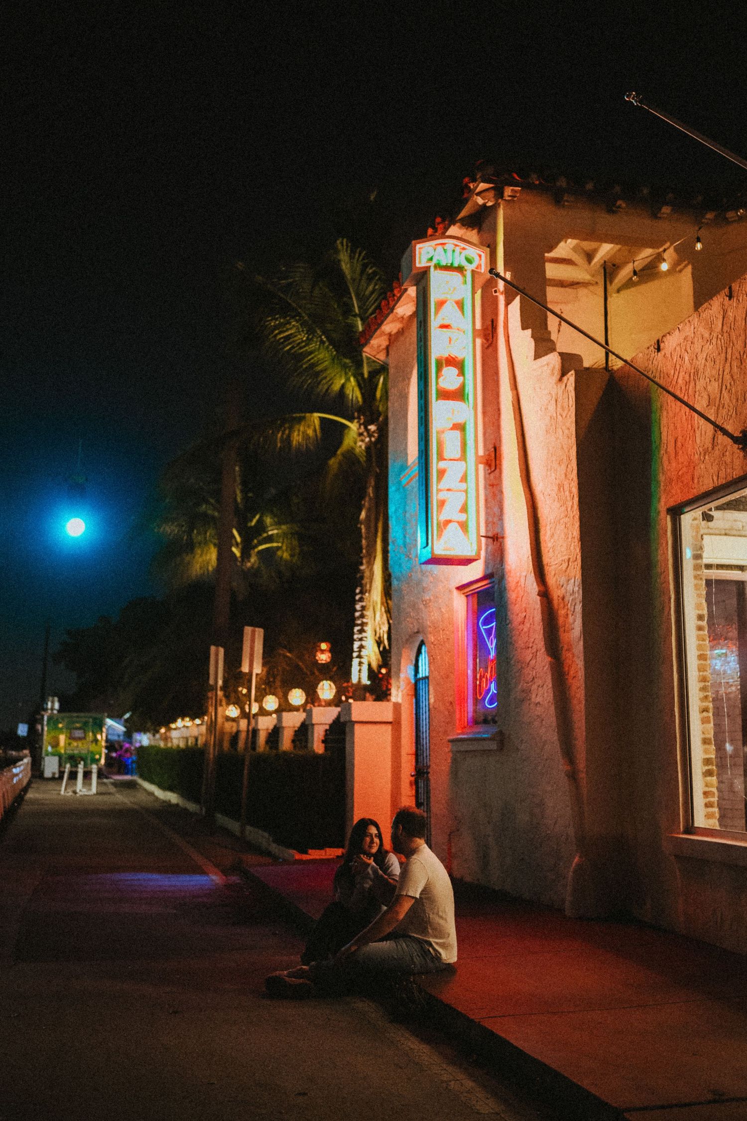 Couple sitting together on a sidewalk at night beside a neon-lit Art Deco building with palm trees under a full moon.
