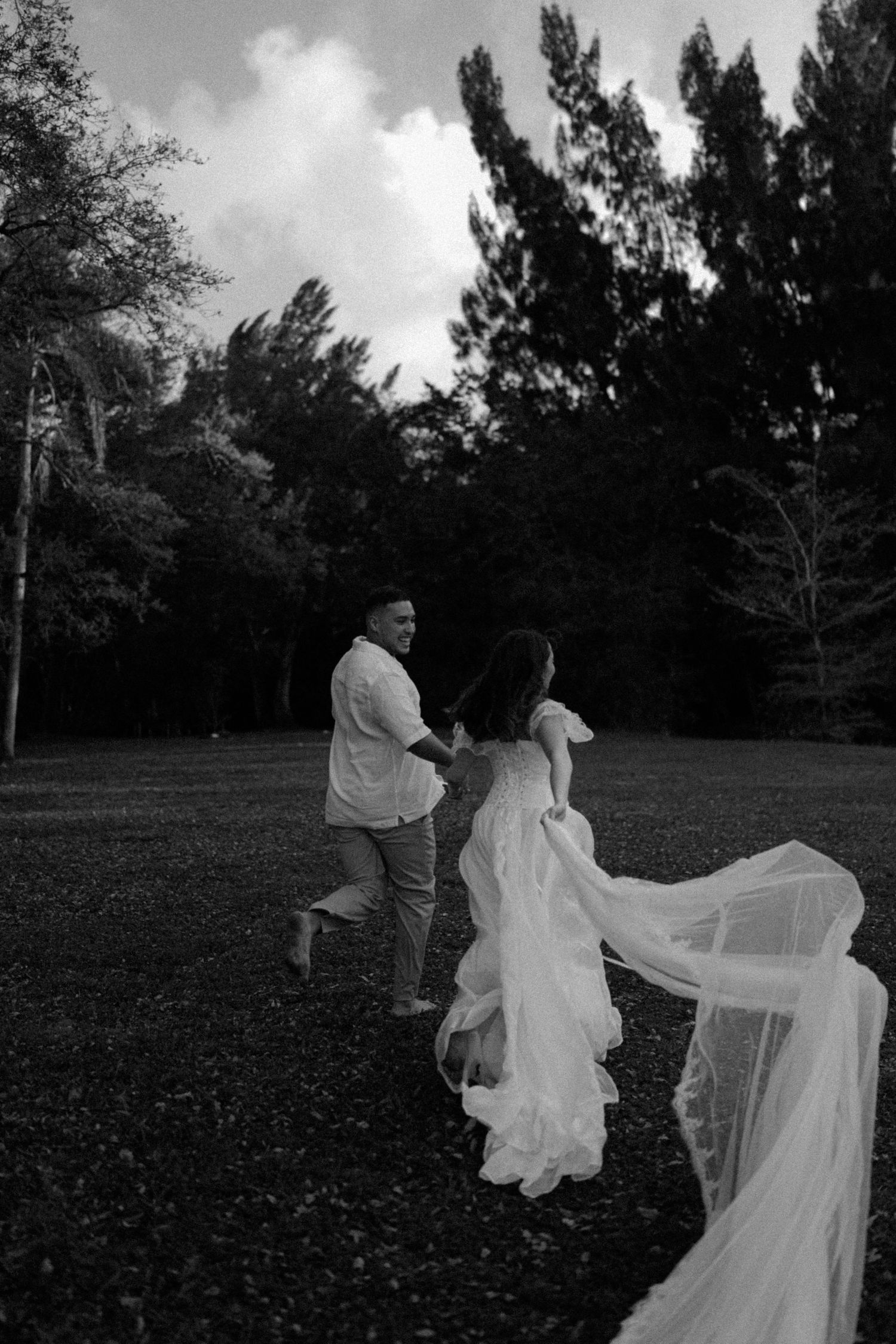 Black and white photo of a bride and groom running through a park, her wedding veil flowing behind them.