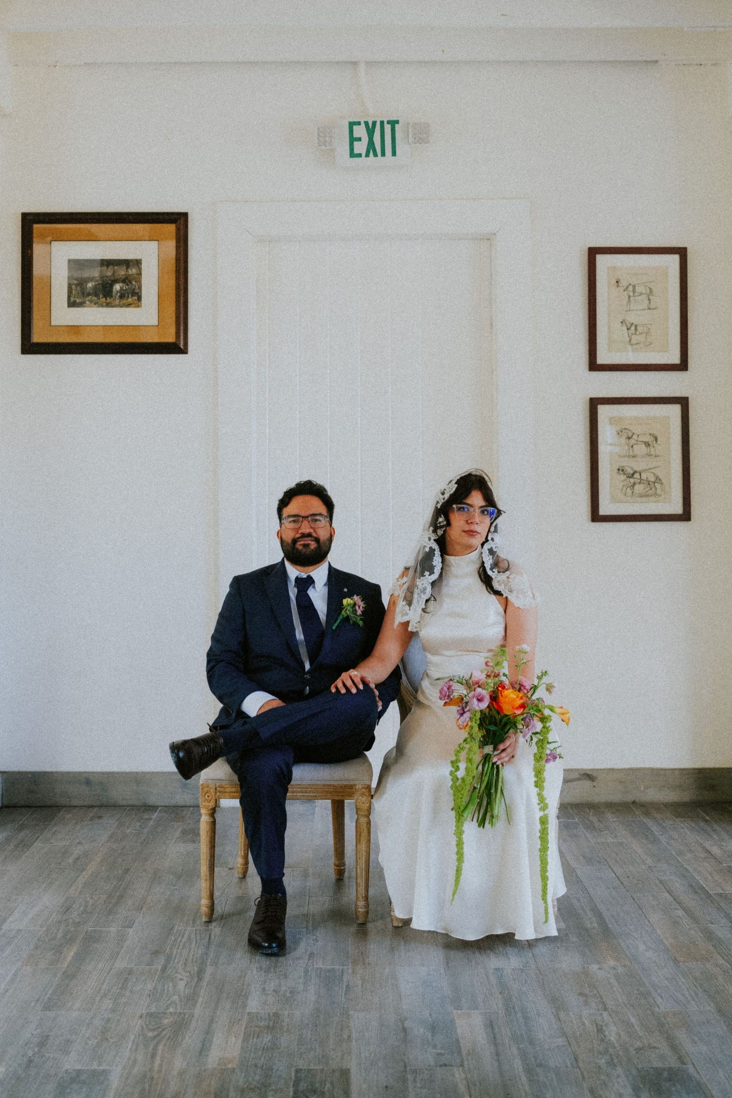 A bride and groom sit side by side in chairs against a white wall, she holds a colorful bouquet.