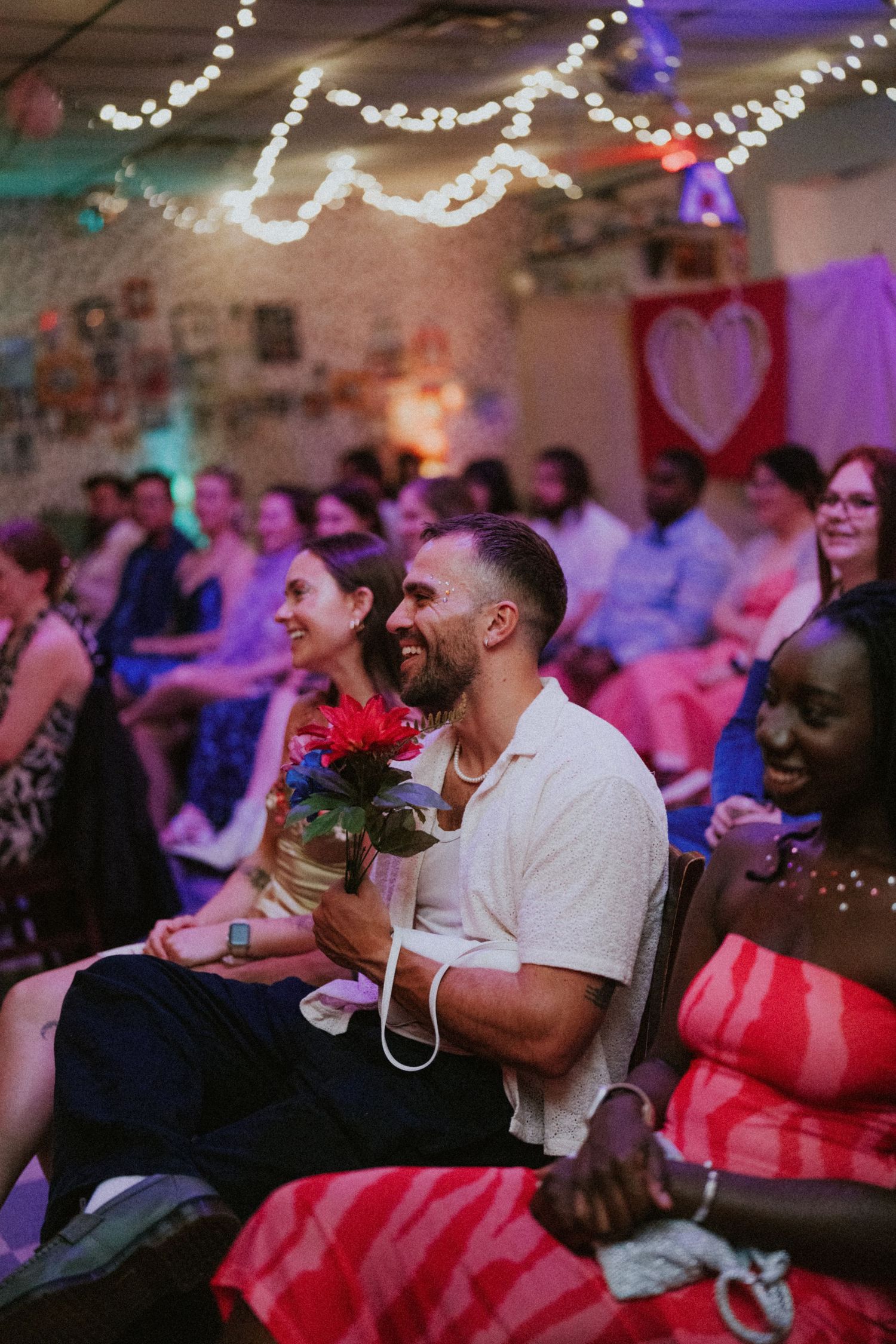 Smiling man holds colorful flowers at a festive indoor event with string lights and a heart banner.