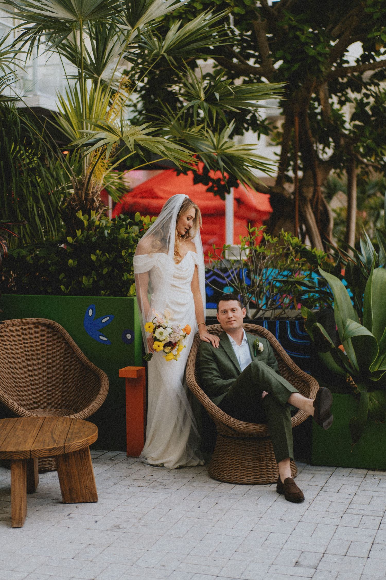 Bride in off-shoulder gown holds bouquet beside groom in green suit among tropical garden decor.