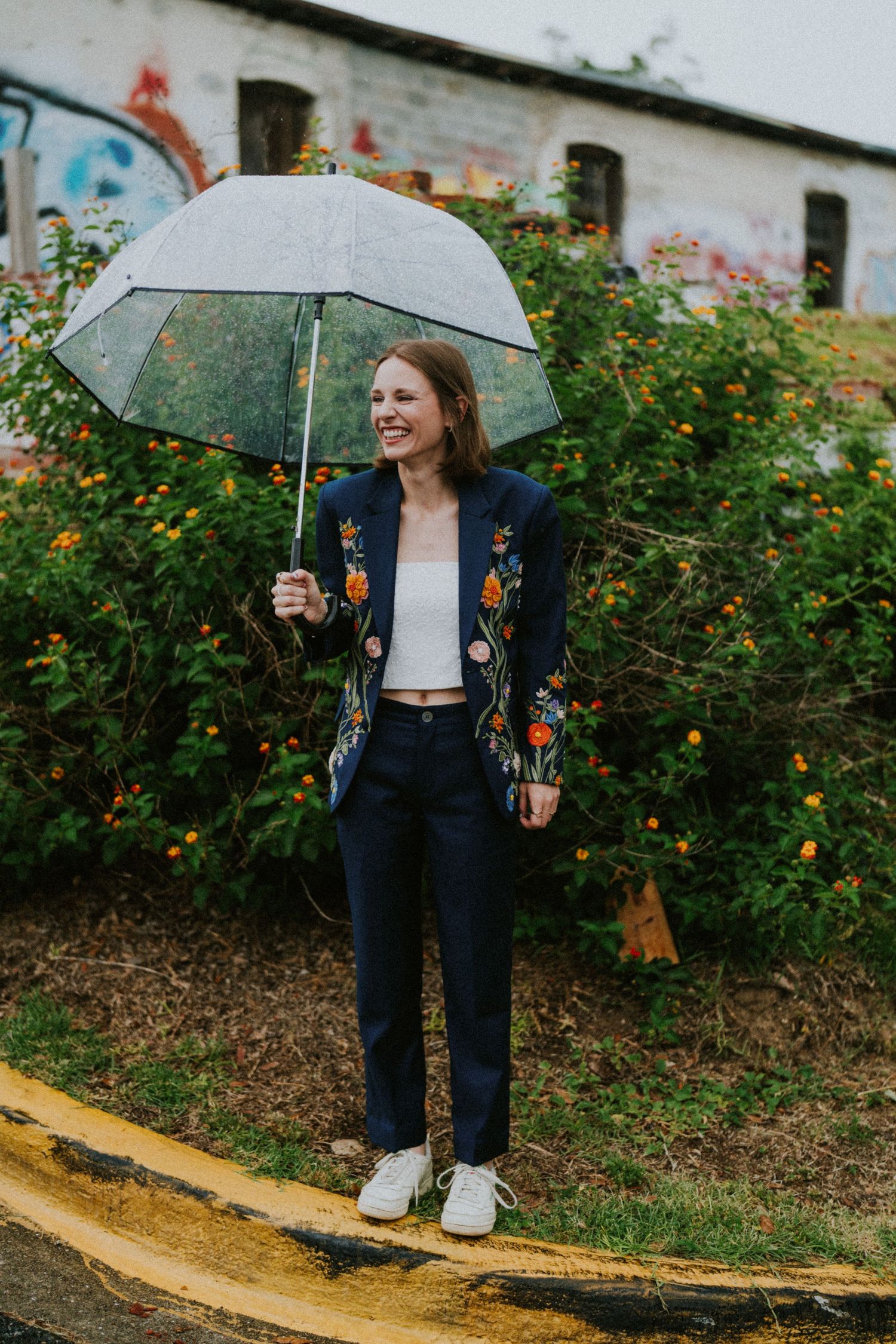 Smiling woman holding clear umbrella in navy floral blazer and trousers, standing outdoors near colorful flowers.