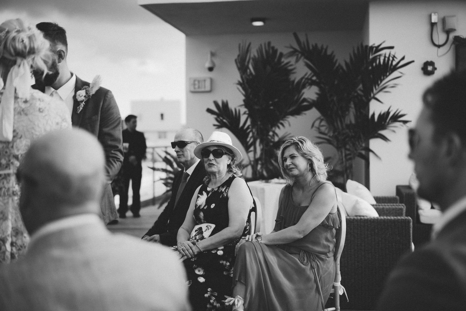 Guests seated outdoors at a wedding ceremony in black and white, including an elegant woman in a floral dress and hat.